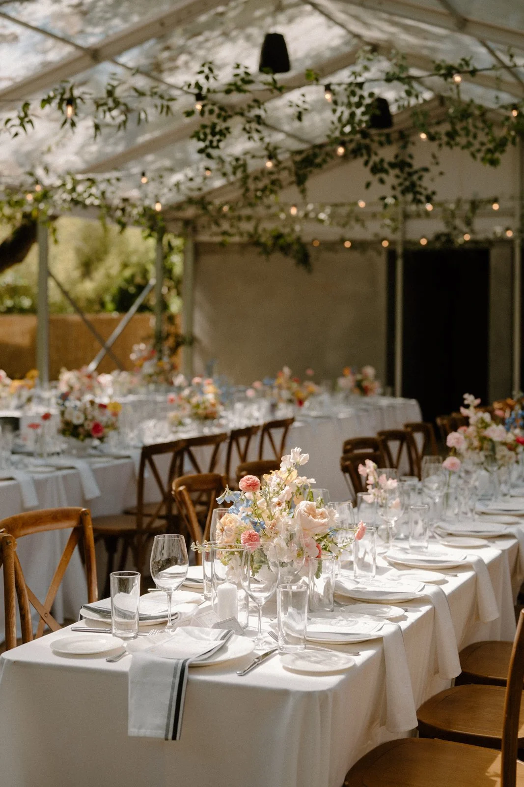 Decorated outdoor event tent with tables set for a meal, featuring floral centerpieces, wine glasses, and tableware under string lights and greenery.