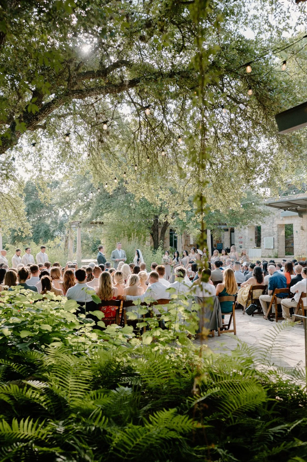 Outdoor wedding ceremony with guests seated and officiant standing in front of the couple, surrounded by trees, string lights, and a stone building in the background.