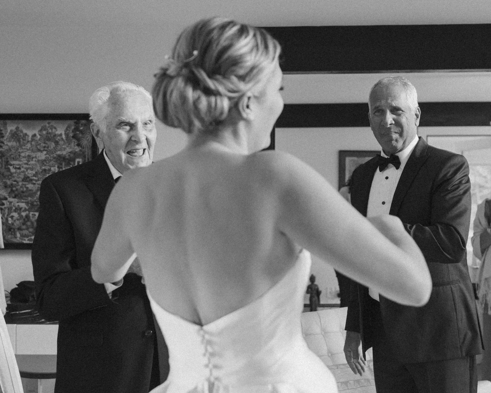 A group of people at a wedding: a woman in a wedding gown, and two men in tuxedos, are visible. The woman is partially turned away, and the men are facing her, smiling.