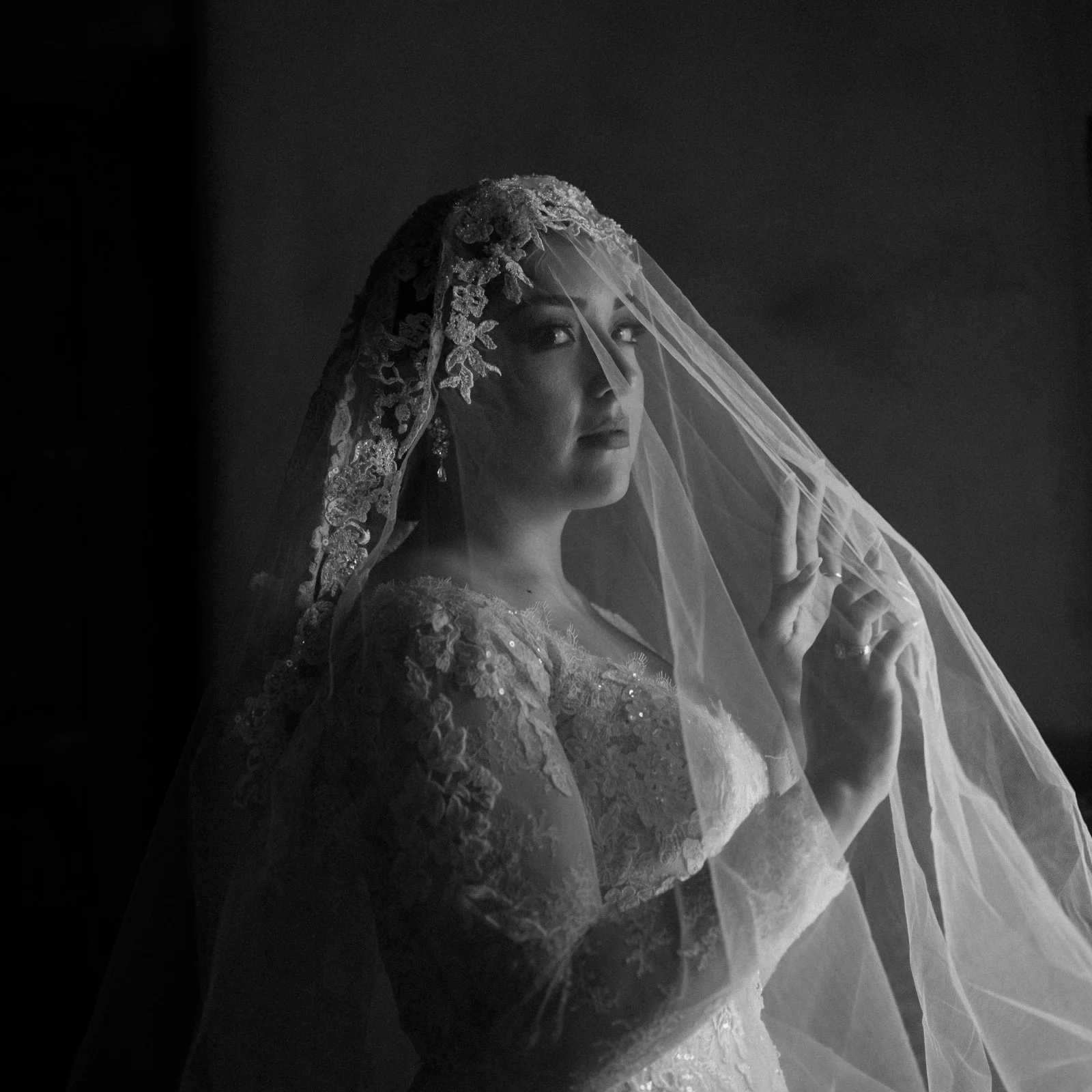 Black and white photo of a woman in a wedding dress with a lace veil, looking over her shoulder with her hand near her face.