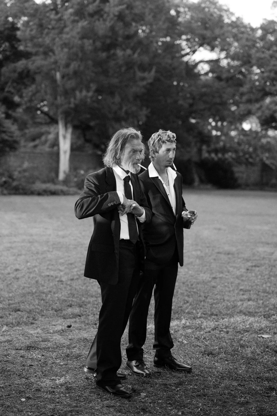 Two men in suits with cigarettes, standing outdoors on a grassy lawn with trees in the background.