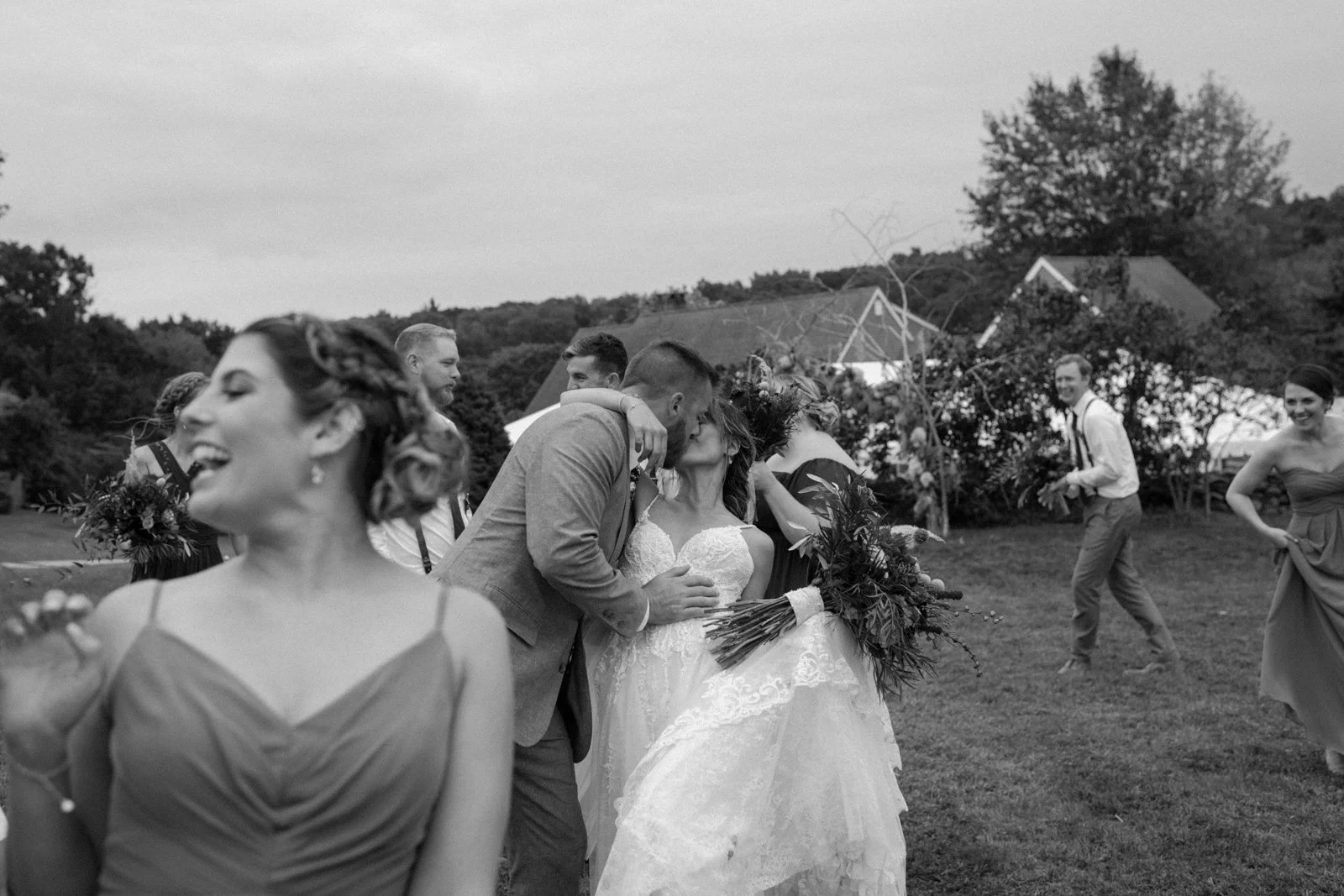 A black-and-white photograph of a wedding celebration outdoors, with a couple kissing in the center surrounded by friends and family.