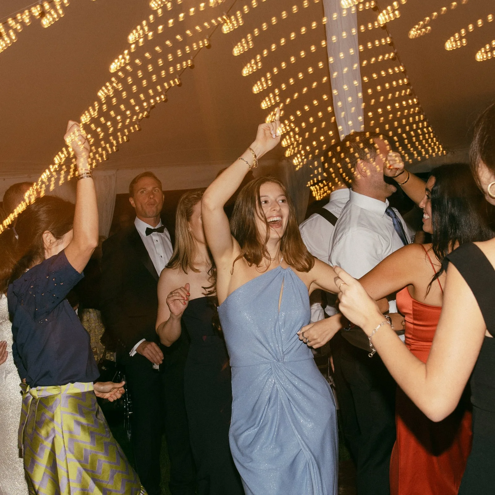 People dancing and celebrating at a party or wedding reception under string lights, with a woman in a blue dress smiling in the center.