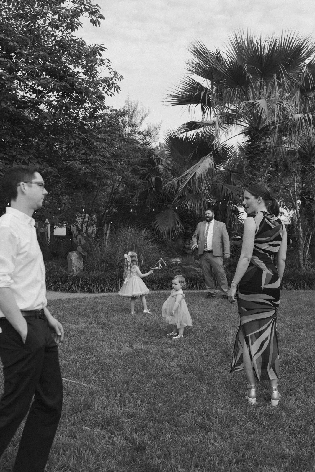 People at an outdoor gathering or party in a lush garden, some children playing and adults engaging, with large palm trees and other foliage in the background.