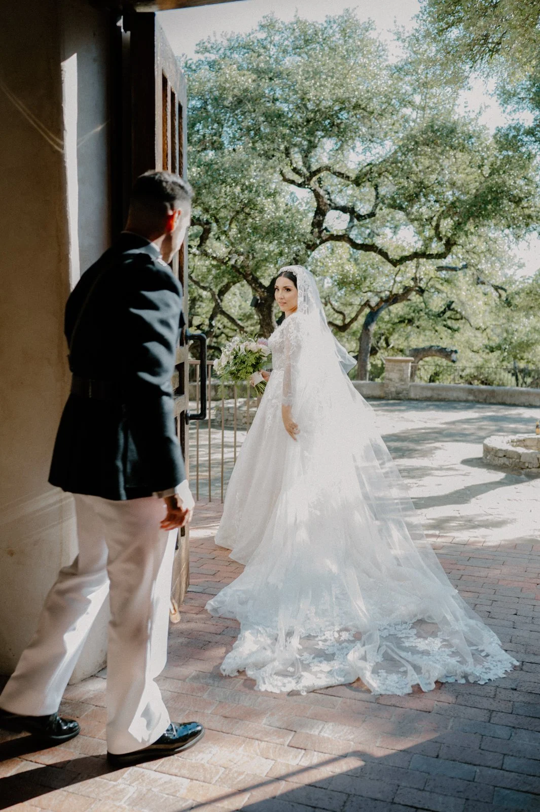 A bride in a white wedding dress holding a bouquet of flowers stands outside, looking back towards a man in formal attire as she steps through a doorway.