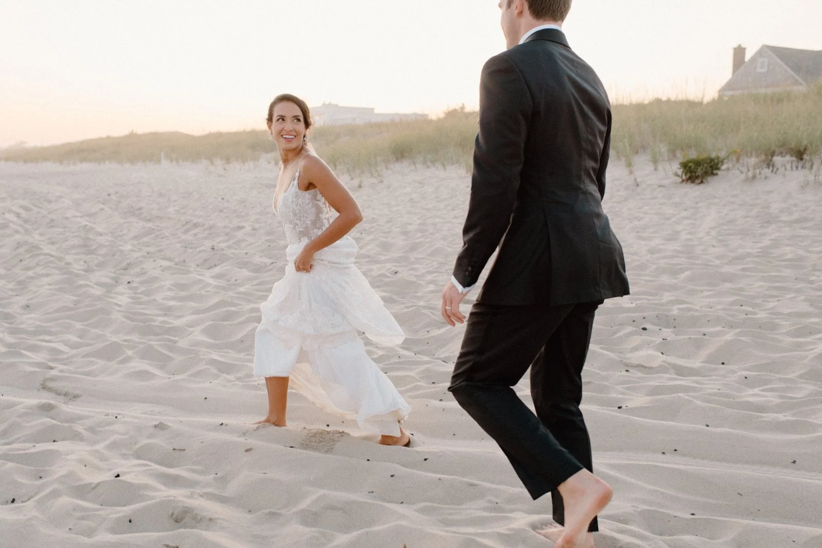 A woman in a white wedding dress walking barefoot on a sandy beach, smiling at a man in a black suit who is also barefoot, with houses and dunes in the background.