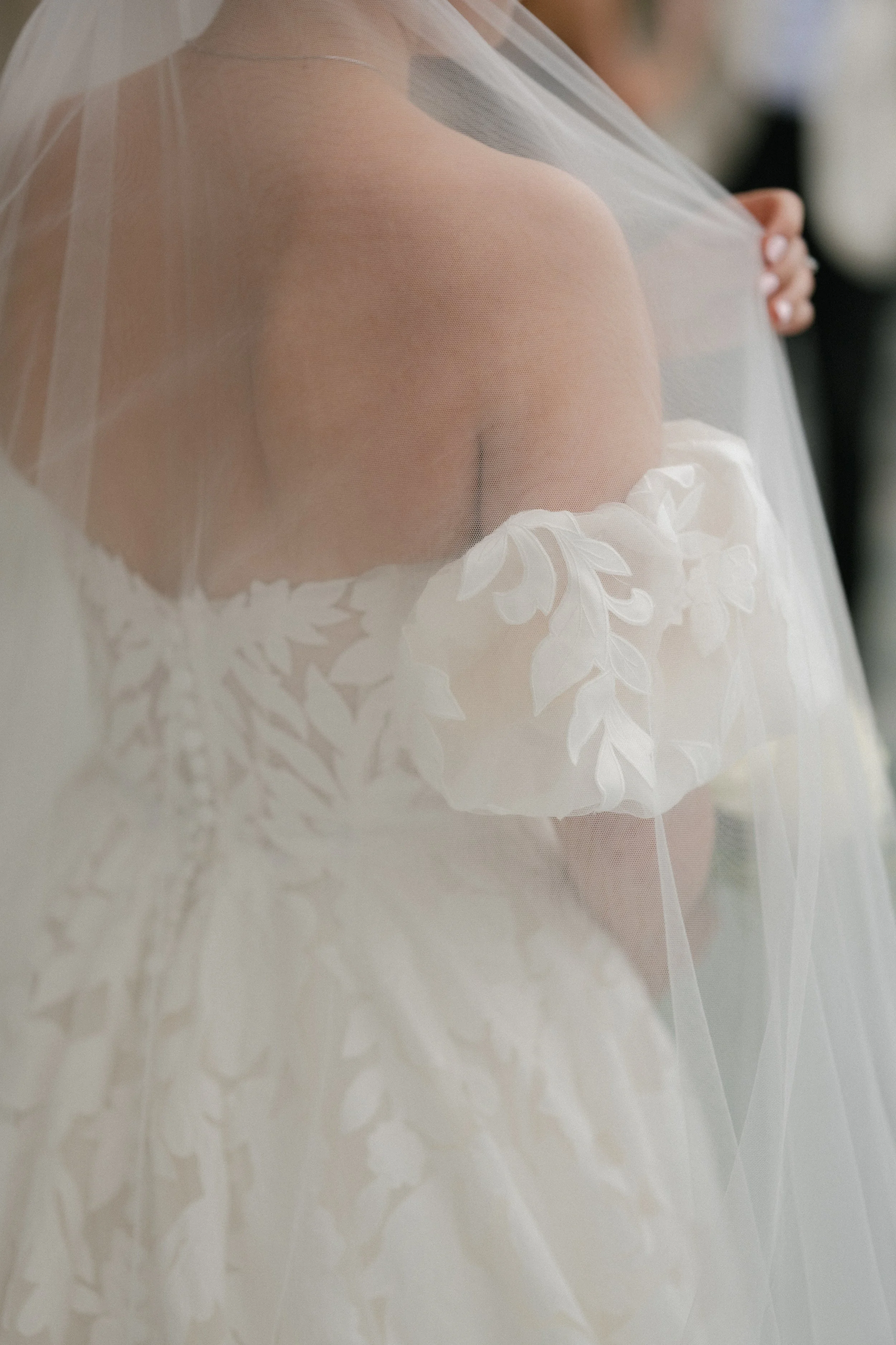 Close-up of a bride wearing a sheer white wedding veil with intricate floral embroidery, holding her hand near her chest.