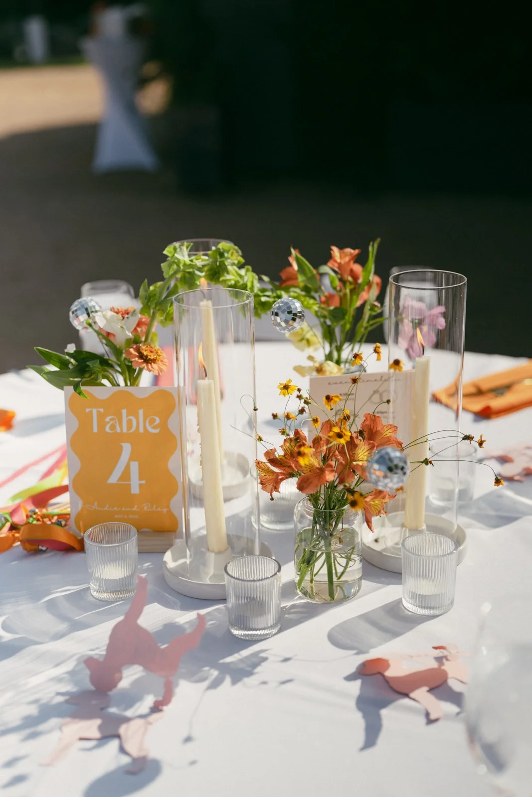 Decorated table with flower arrangements, candles, glassware, a sign indicating "Table 4," and paper confetti, set outdoors for a celebration.