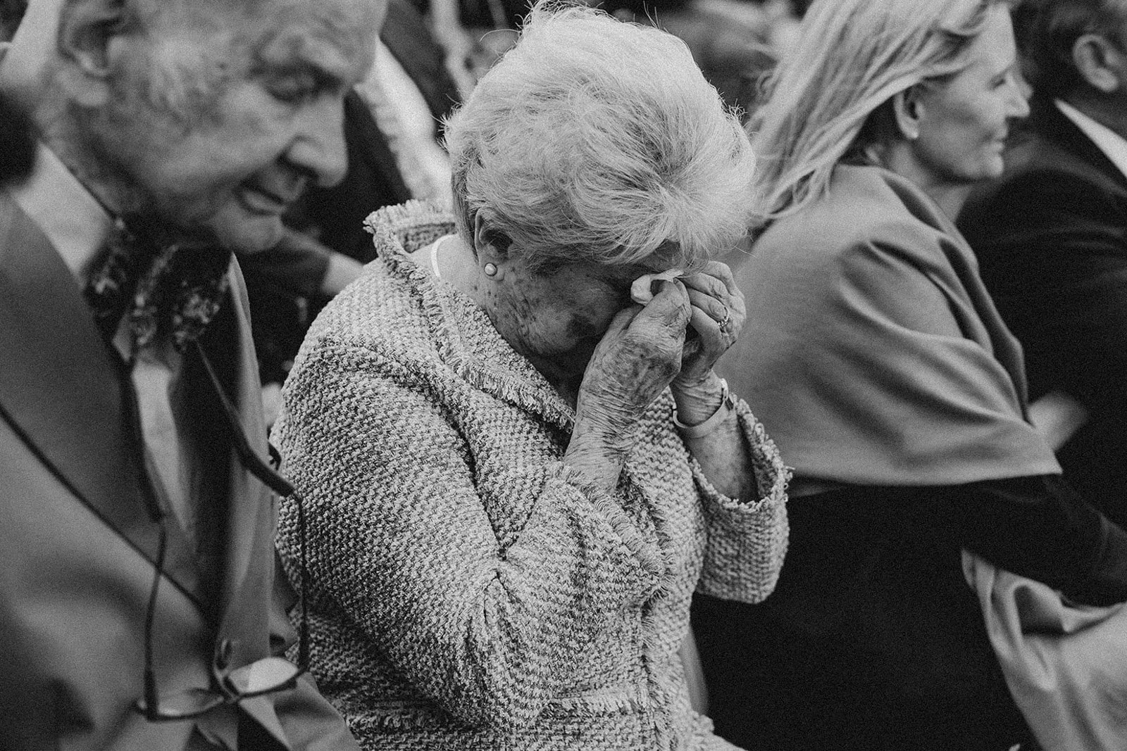 An elderly woman with white hair covering her face with her hands, sitting in a row of people at an event.