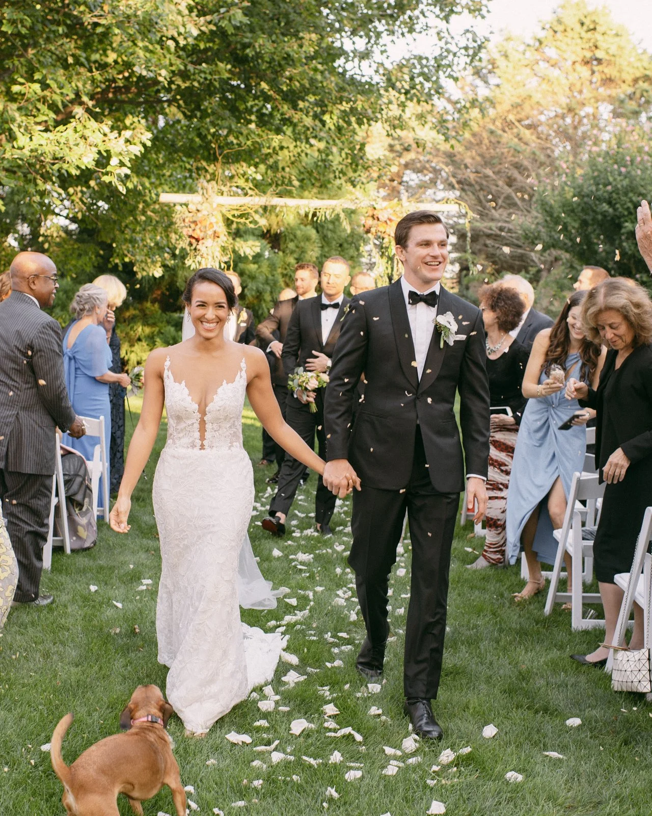 Bride and groom walking hand in hand at their outdoor wedding ceremony, surrounded by guests celebrating with confetti, trees, and a decorated archway in the background, with a dog in the foreground.