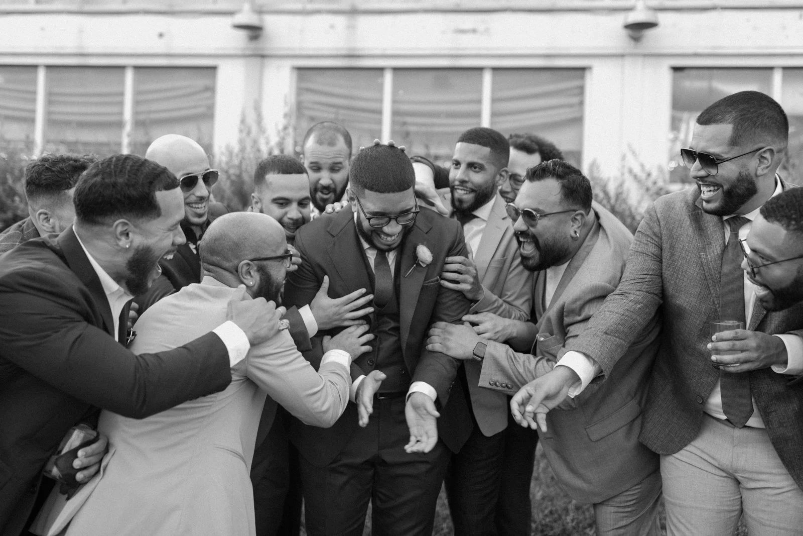 Group of men celebrating and laughing together outdoors during daytime, wearing suits and sunglasses.