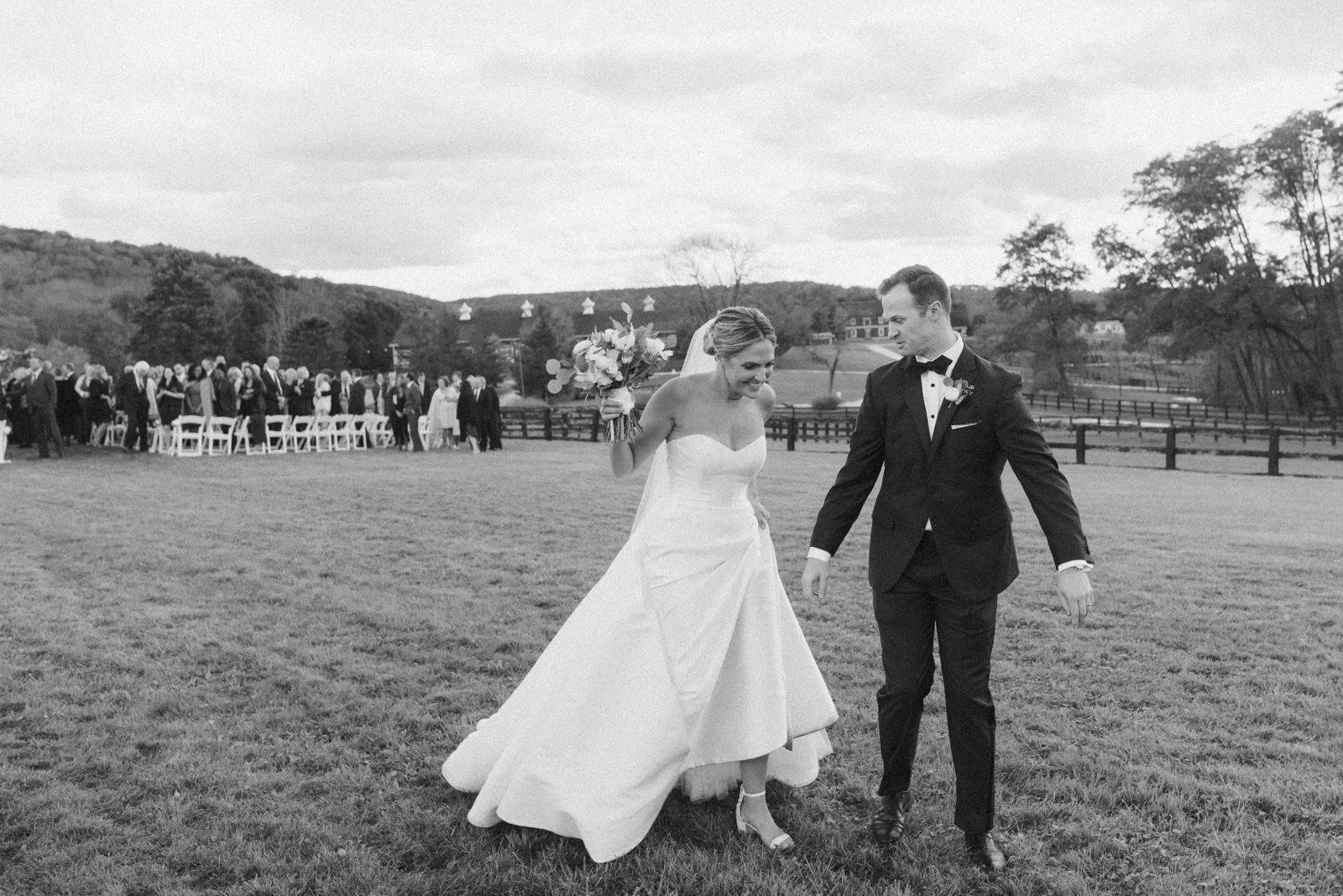 Black and white photo of a bride and groom smiling and walking across a grassy field, with a group of people standing in the background under a cloudy sky.