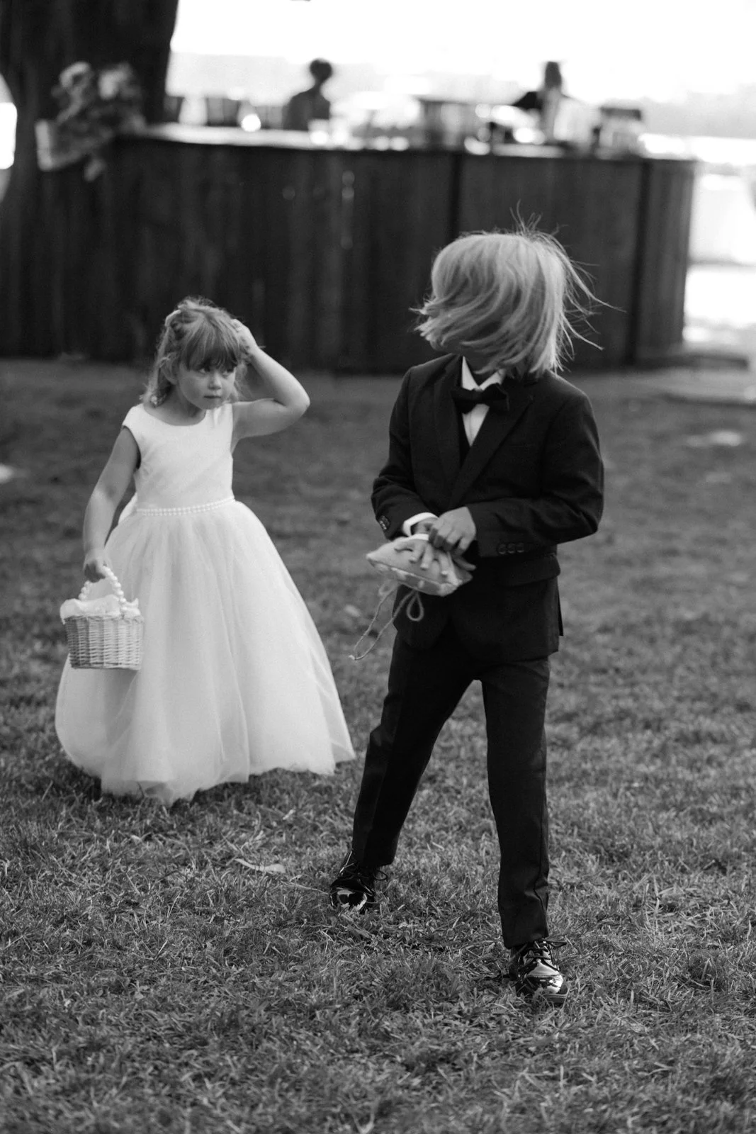A black and white photo of a young girl in a white dress carrying a basket and an older boy in a tuxedo holding a pillow with rings, standing outdoors on grass during a wedding ceremony.