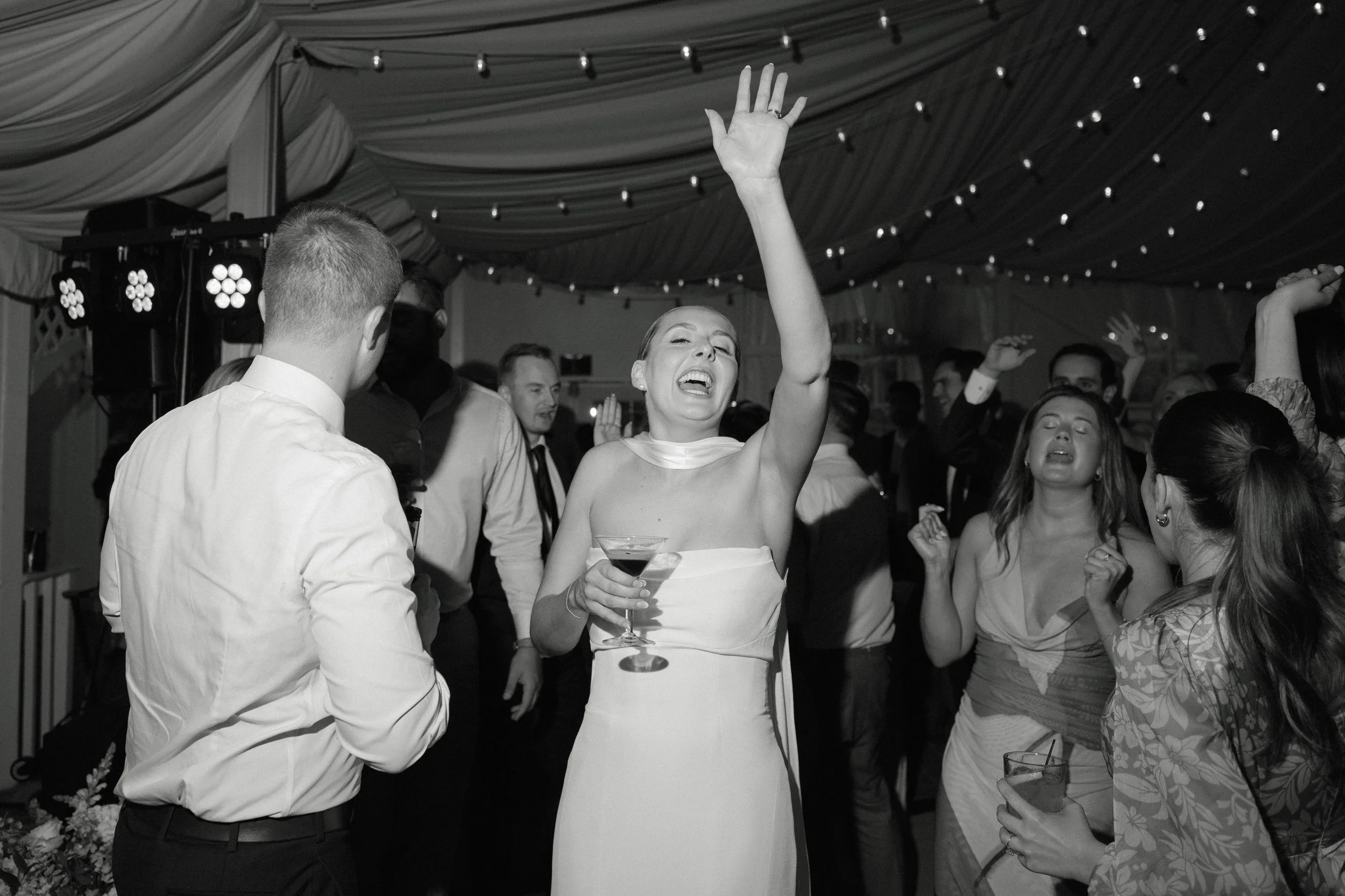 A black and white photo of a lively celebration with people dancing and socializing under a decorated tent. The focus is on a woman in a strapless dress holding a cocktail and raising her hand, with other guests around her enjoying the event.