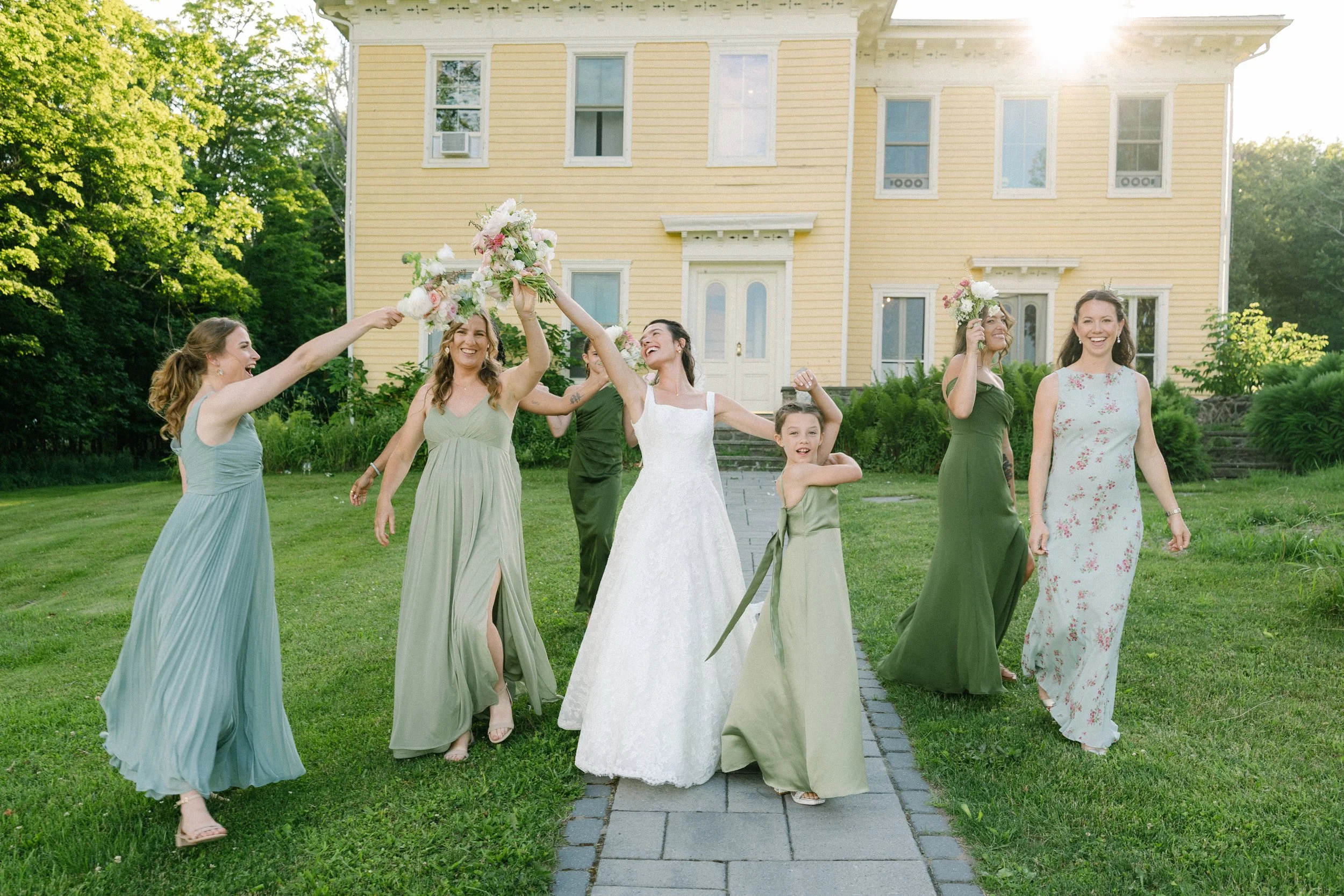 Women in colorful dresses celebrating outdoors on a lush green lawn in front of a yellow house with white trim, during what appears to be a wedding or special event, with sunlight shining from the upper right corner.
