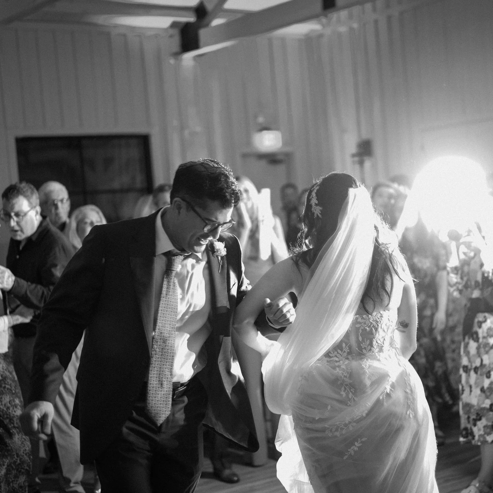 A black and white photo of a wedding reception showing a man in a suit and glasses dancing with a woman in a wedding dress with a veil. Other guests are visible in the background.