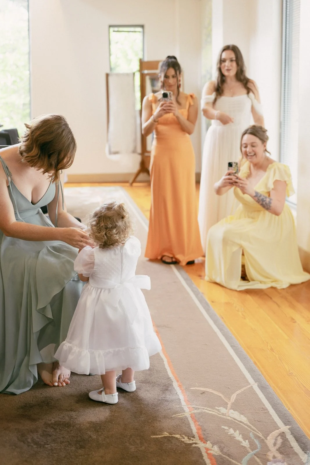 Women and children preparing for a special occasion in a bright, airy room with large windows, some taking photos and a young girl in a white dress with an adult woman in a gray dress and a girl in a yellow dress with tattoos.