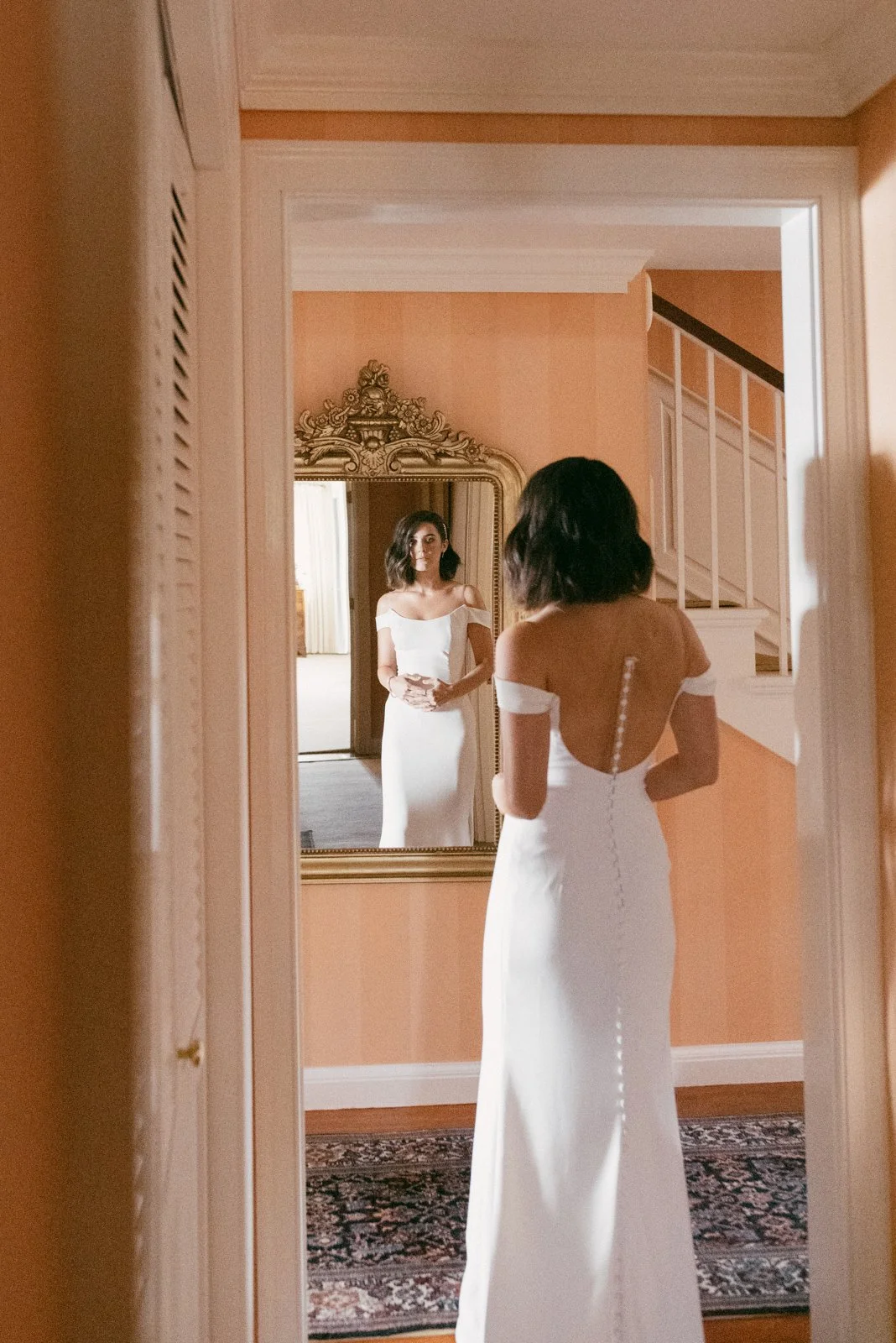 Woman in a white wedding dress looking at herself in a mirror, standing in a warmly lit room with peach-colored walls.