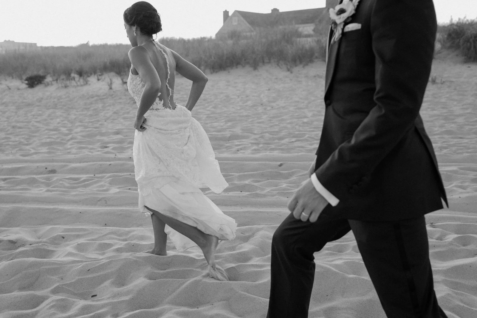 A bride in a wedding dress walking barefoot on sand with a groom in a suit nearby at a beach, with a house and dunes in the background.