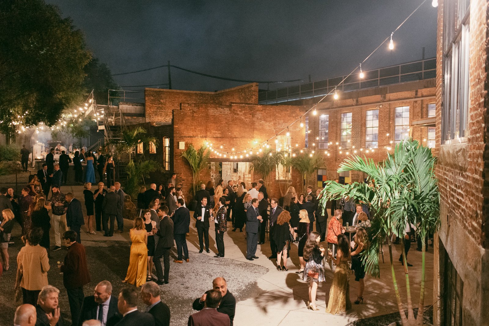 Nighttime outdoor social event with people in formal attire, string lights, brick buildings, and tropical plants.