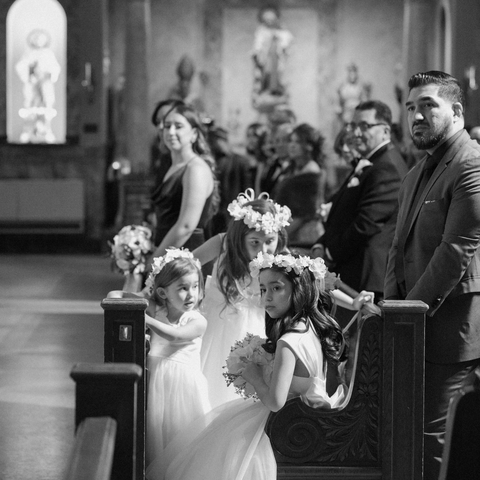 Children and adults dressed in formal attire at a wedding ceremony inside a church with religious statues on the wall, some children wearing flower crowns, and the bride holding a bouquet.