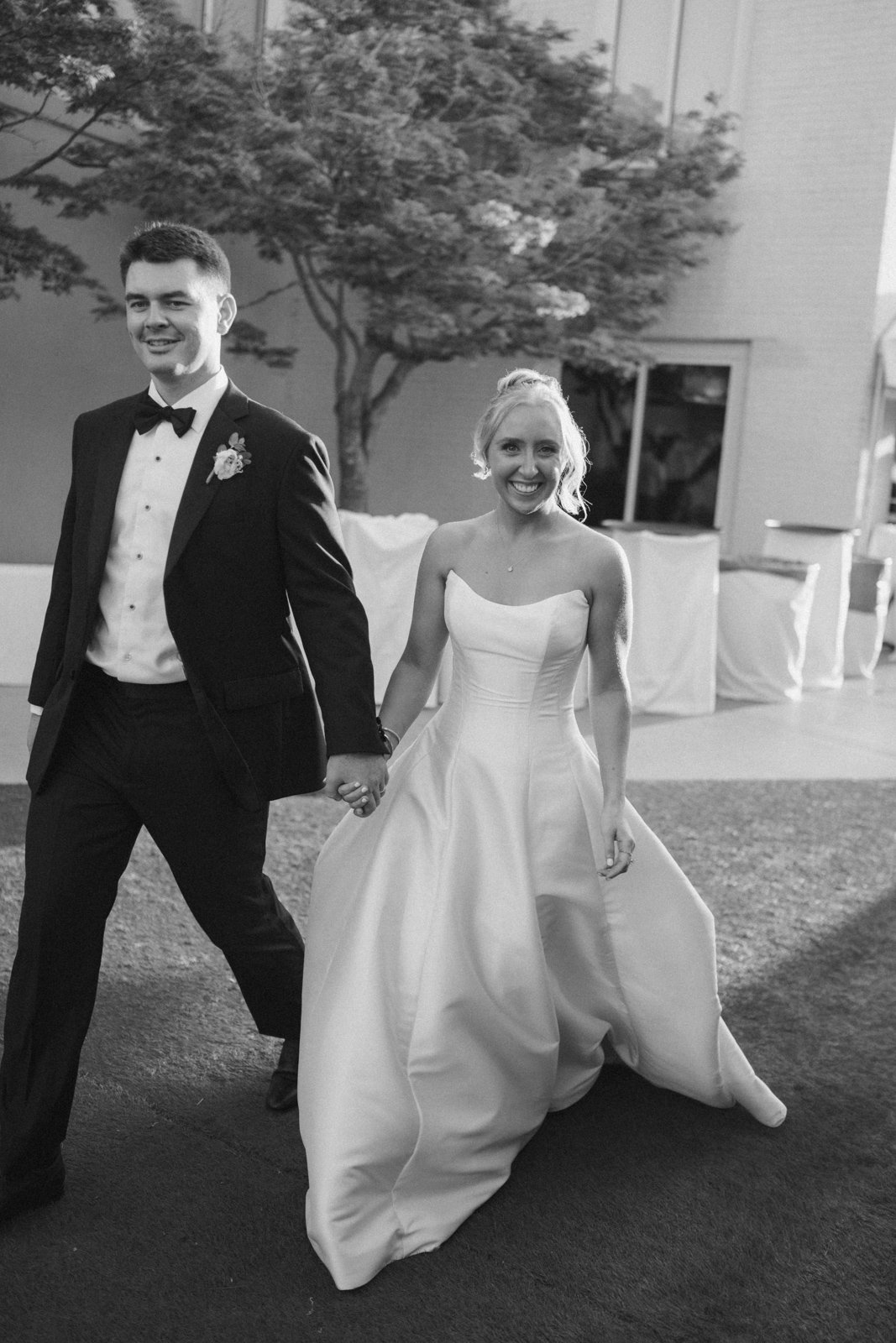 A bride and groom holding hands and walking outdoors at sunset, dressed in wedding attire, smiling, with trees and wedding decorations in the background.
