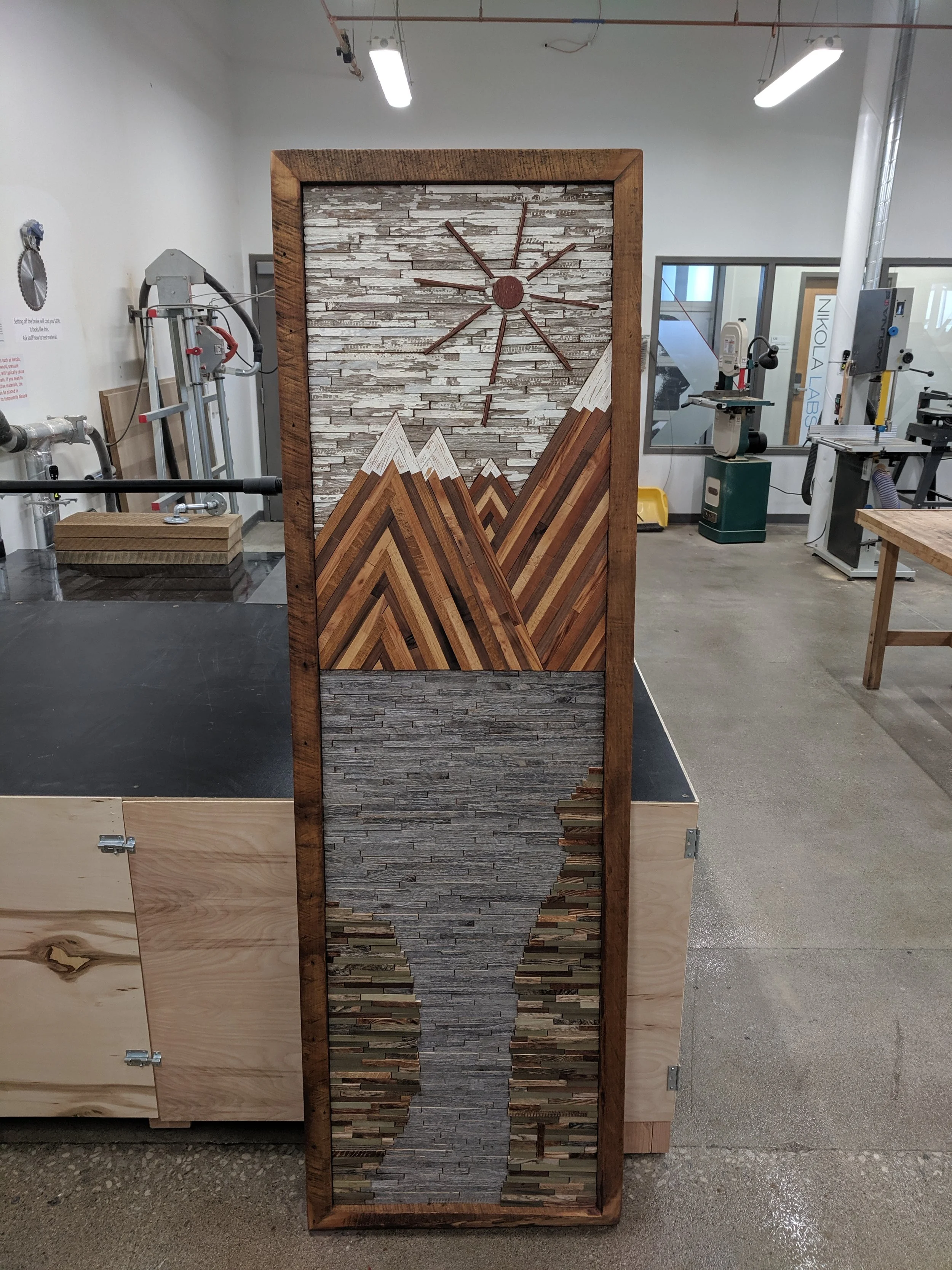 A tall wooden frame displays a mosaic wall with three sections: top section has white and gray stacked stone, middle section has wood and stone arranged in a pointed mountain pattern, and bottom section features dark and light wood pieces.