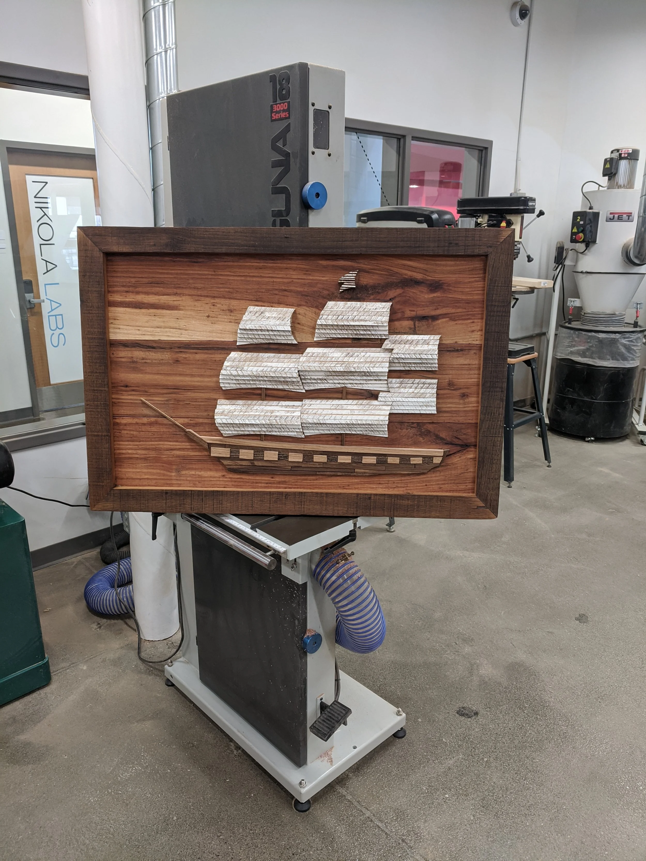 A woodworking or crafting setup with a framed piece of furniture in progress on a machine, surrounded by various tools and machinery in a workshop.