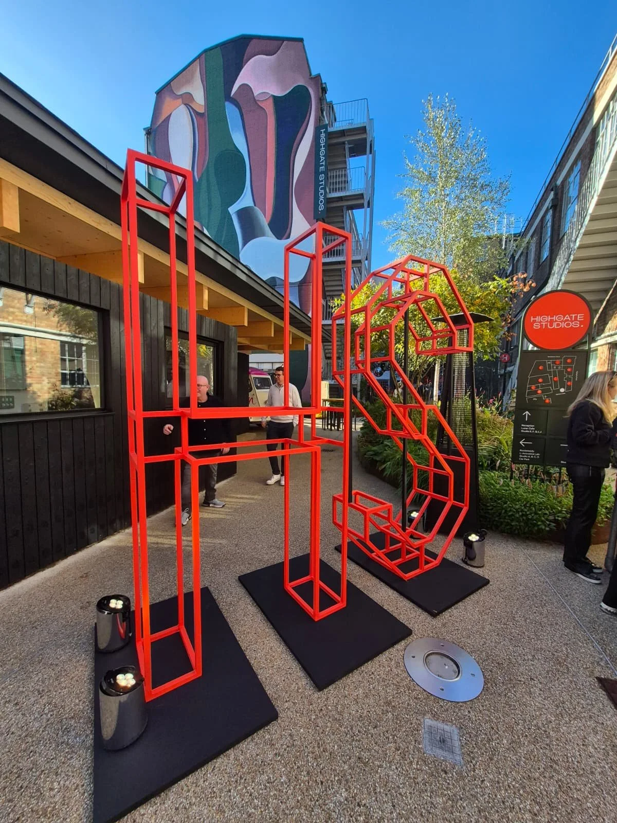 Outdoor art installation with red geometric metal structures on black bases, set in a public space. In the background, there is a black building with large windows, a mural on a tall building, and people walking around. The scene is sunny with a clear blue sky and trees.