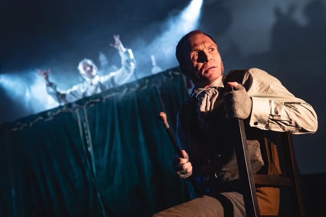 A man with a distressed expression, dressed in old-fashioned clothing, holding a cane and sitting on a wooden chair, with a puppet of a person with outstretched hands in the background on a puppet stage.