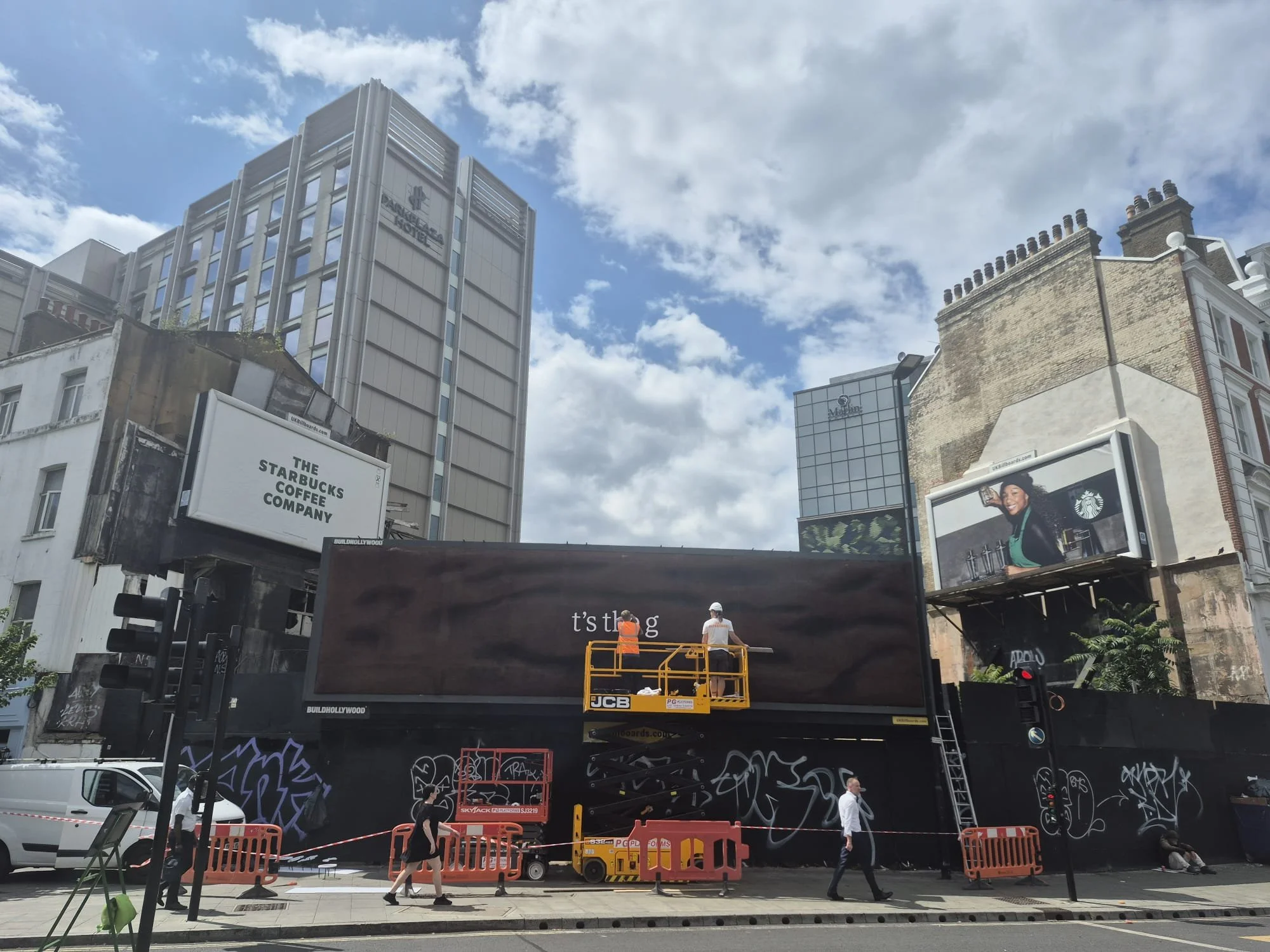 Construction workers are installing a large billboard in an urban area. The billboard displays an advertisement with a woman holding coffee cups, promoting Starbucks. Several buildings and billboards can be seen in the background.