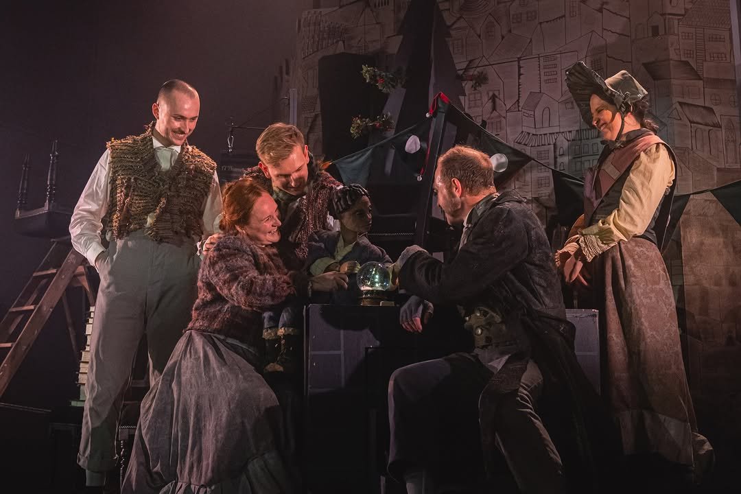 A theatrical stage scene with six actors gathered around a piano, some leaning in and smiling, in period costumes, with a painted cityscape backdrop.