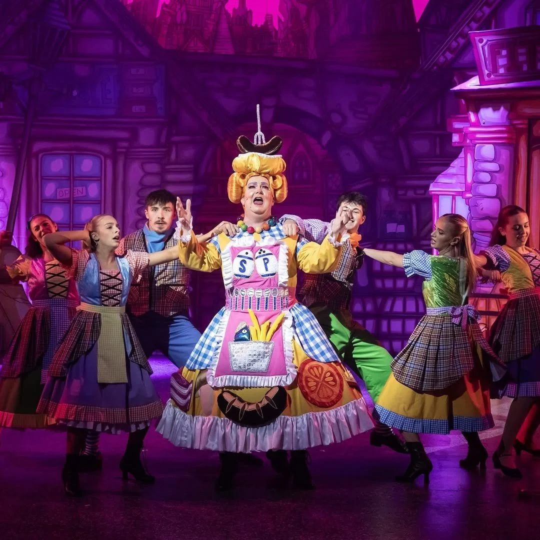 The stage features a theatrical performance with a woman in a colorful costume as the central figure, surrounded by young performers in matching vibrant costumes. The background depicts a whimsical, cartoon-like cityscape in purple hues.