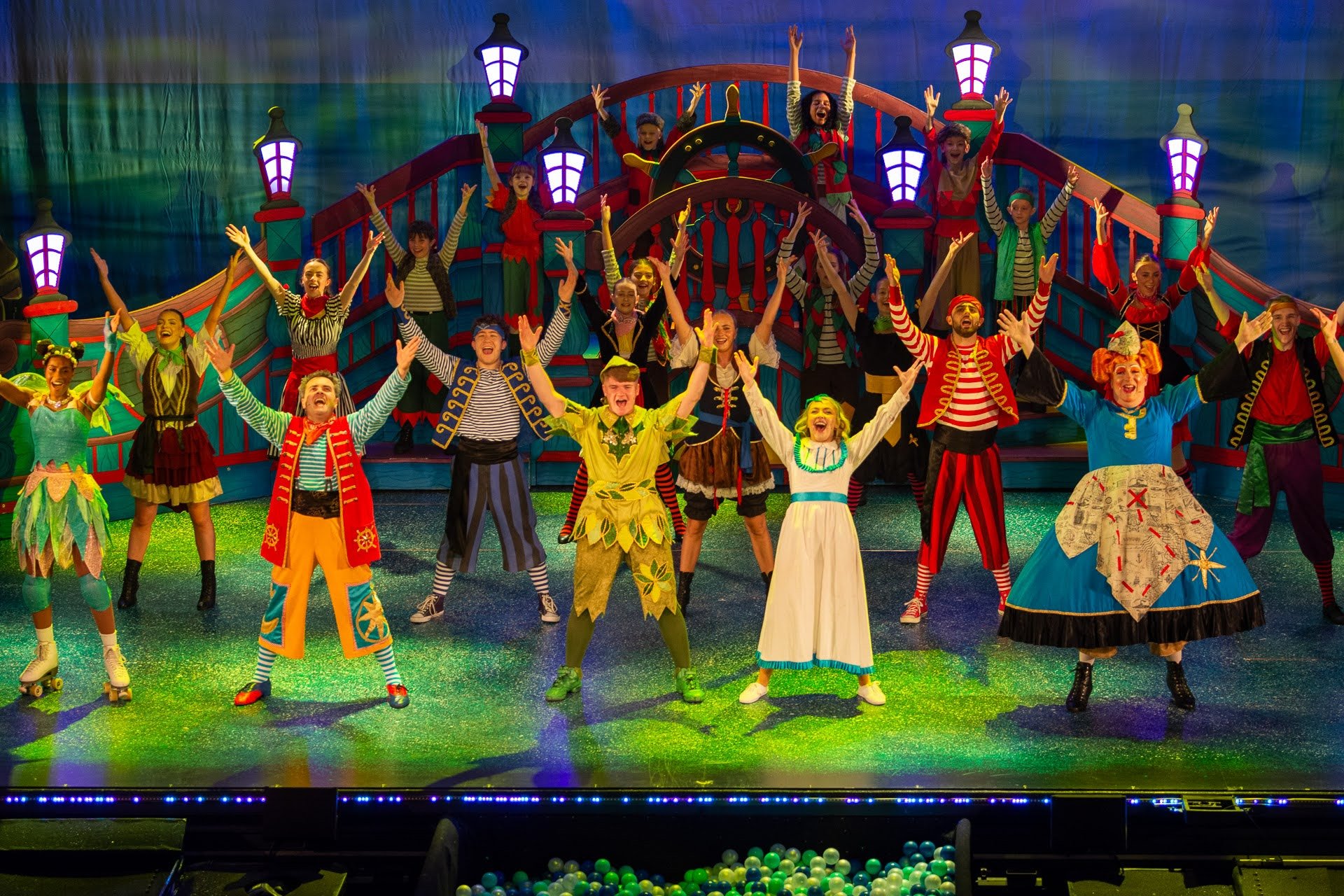 A colorful stage production with children and adults dressed in vibrant costumes, performing a musical or play with a nautical theme, featuring a ship set in the background.
