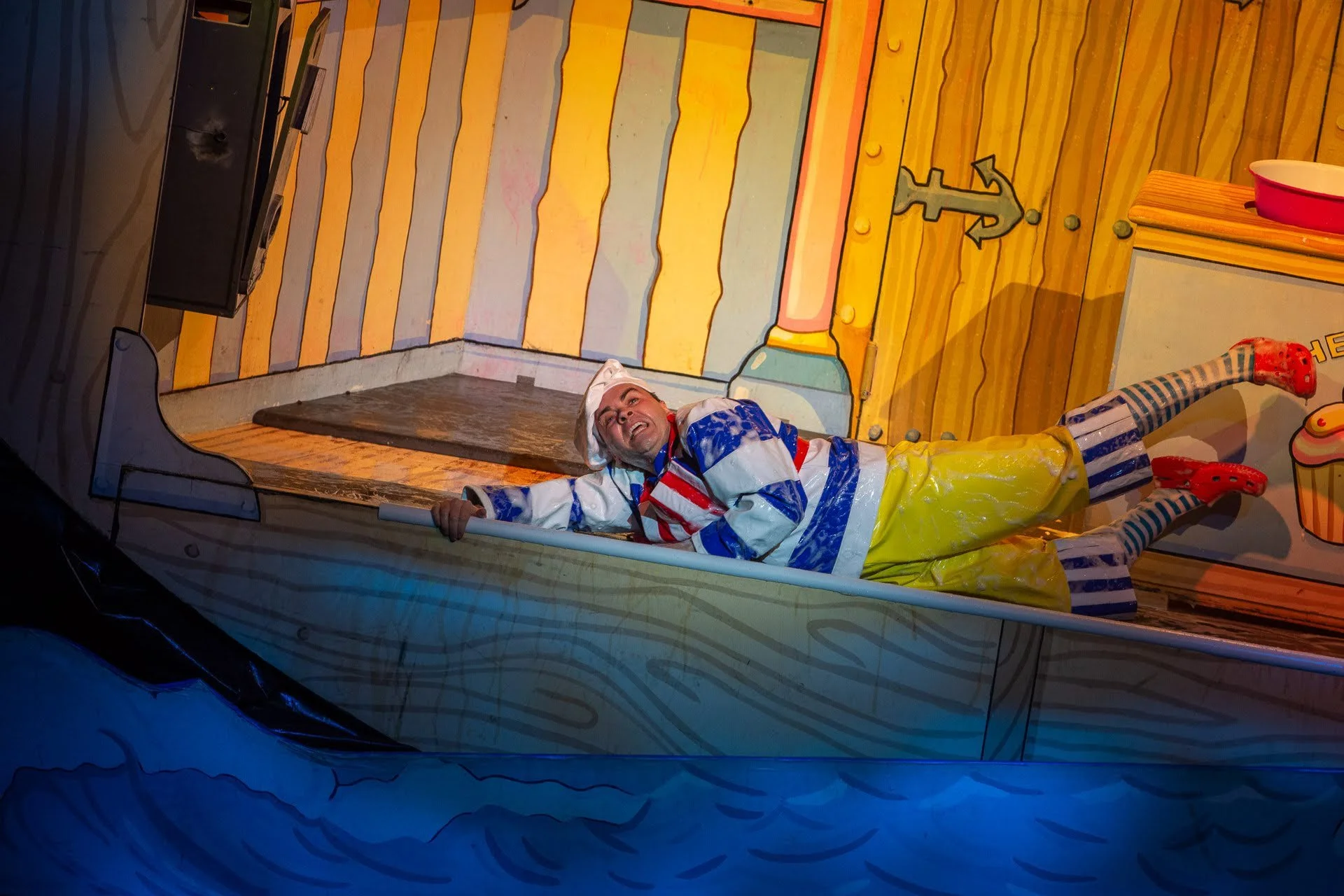 Person lying in a colorful boat cake with a painted wooden panel background, dressed in clown-themed clothing and a white hat.