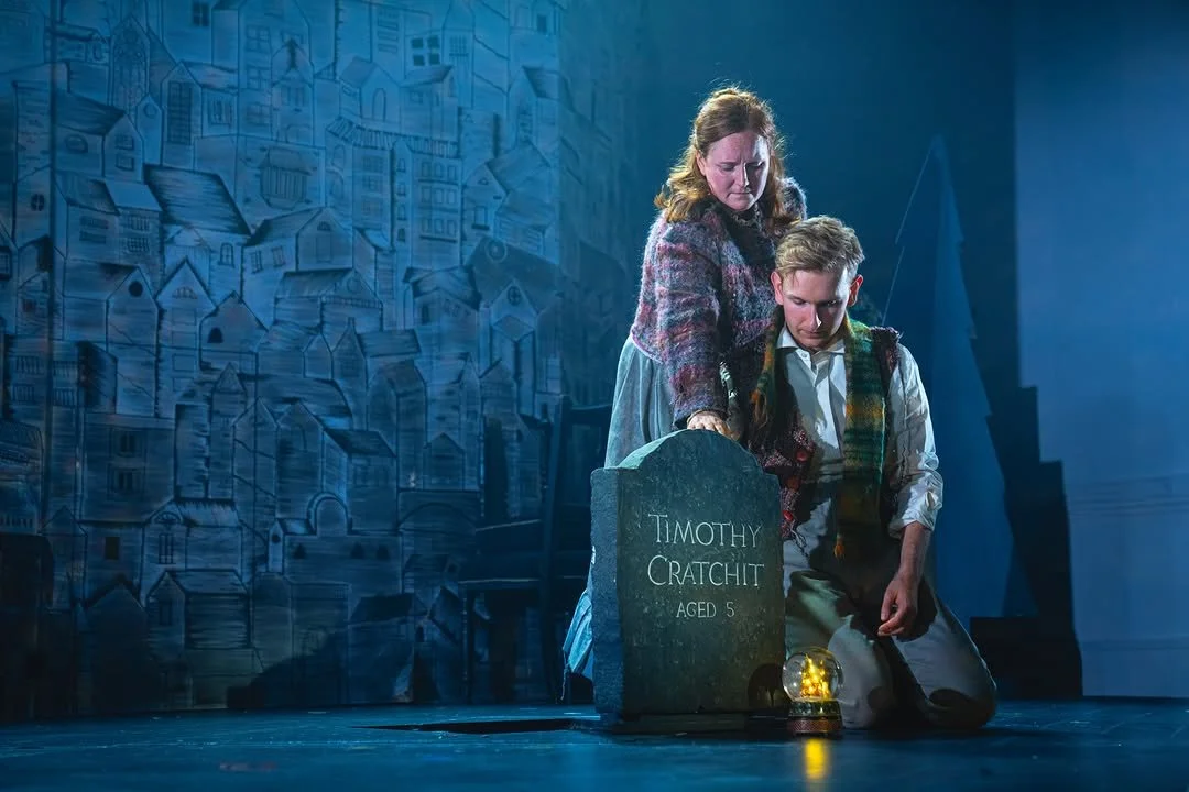 A woman and a boy kneeling next to a baby grave with the name Timothy Cratchit, aged 5, inscribed on it. The woman is touching the gravestone. There is a small lantern next to the gravestone.