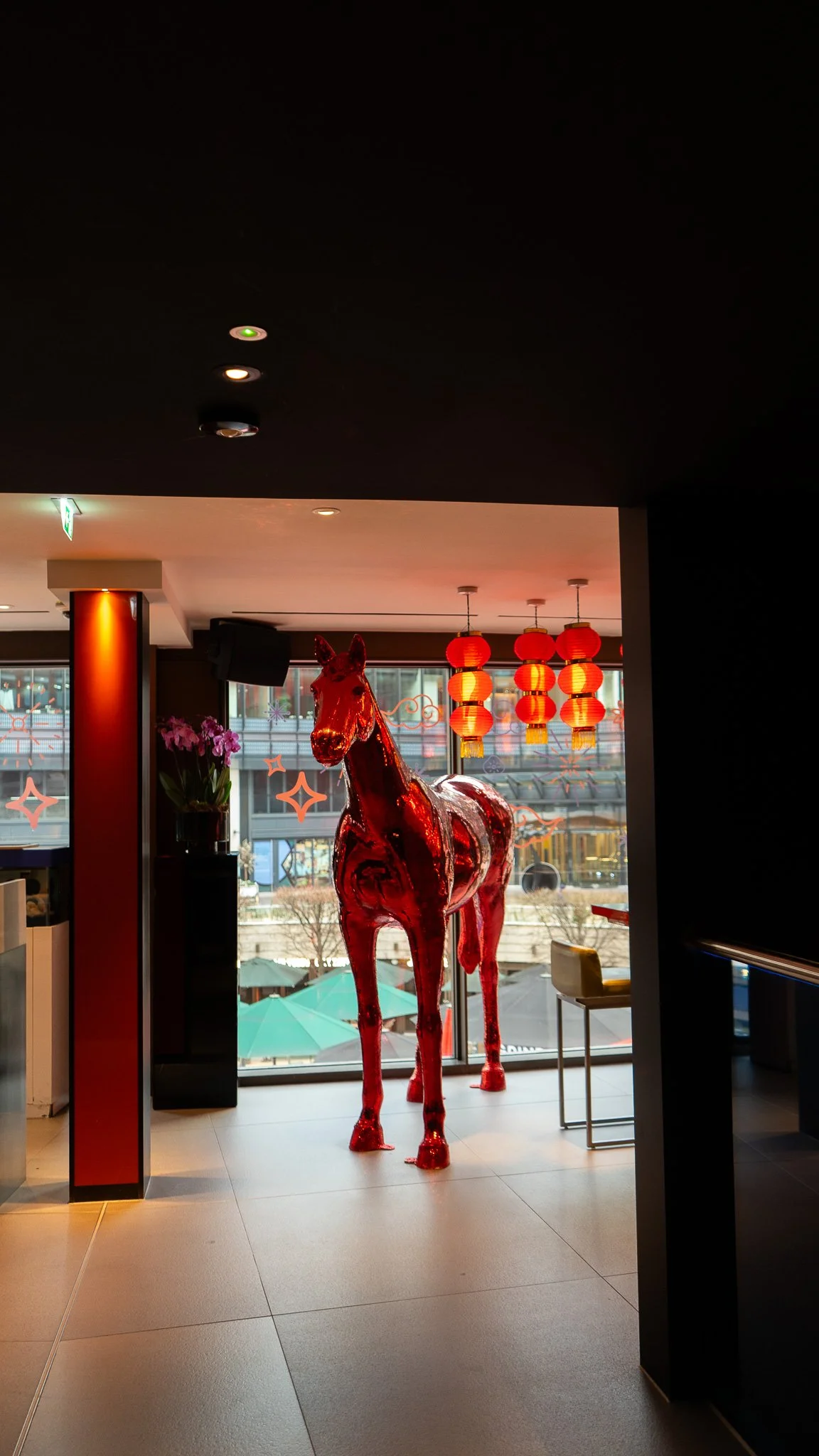 Red metallic horse sculpture inside a room with large windows, hanging red lanterns, and a potted plant in the background.