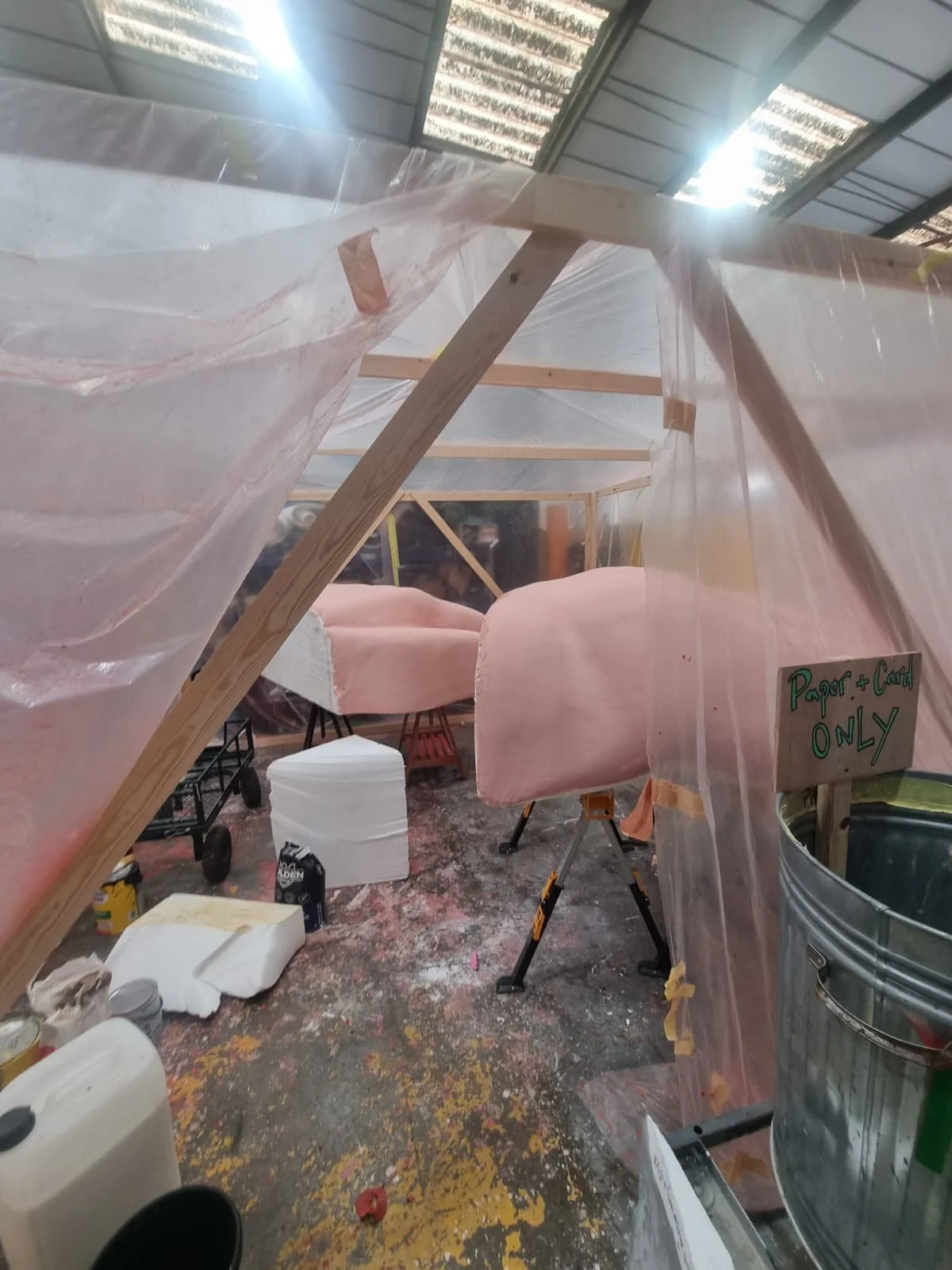Inside a workshop with beams and plastic sheeting, pink foam or insulation panels, tools, and materials scattered around. A sign says 'Paper + Card ONLY' on a metal trash can.