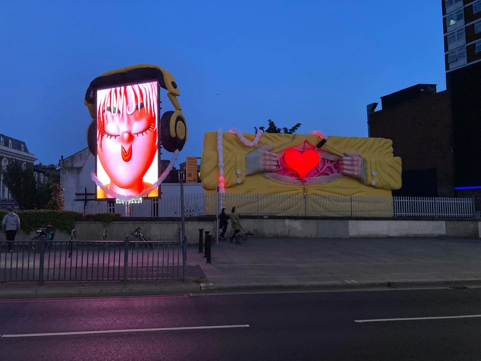 Urban scene at dusk with a digital billboard displaying a cartoon face wearing headphones and pink dripping hair, and a large mural of a person unzipping a yellow jacket to reveal a glowing red heart, with people walking and a dog in front.
