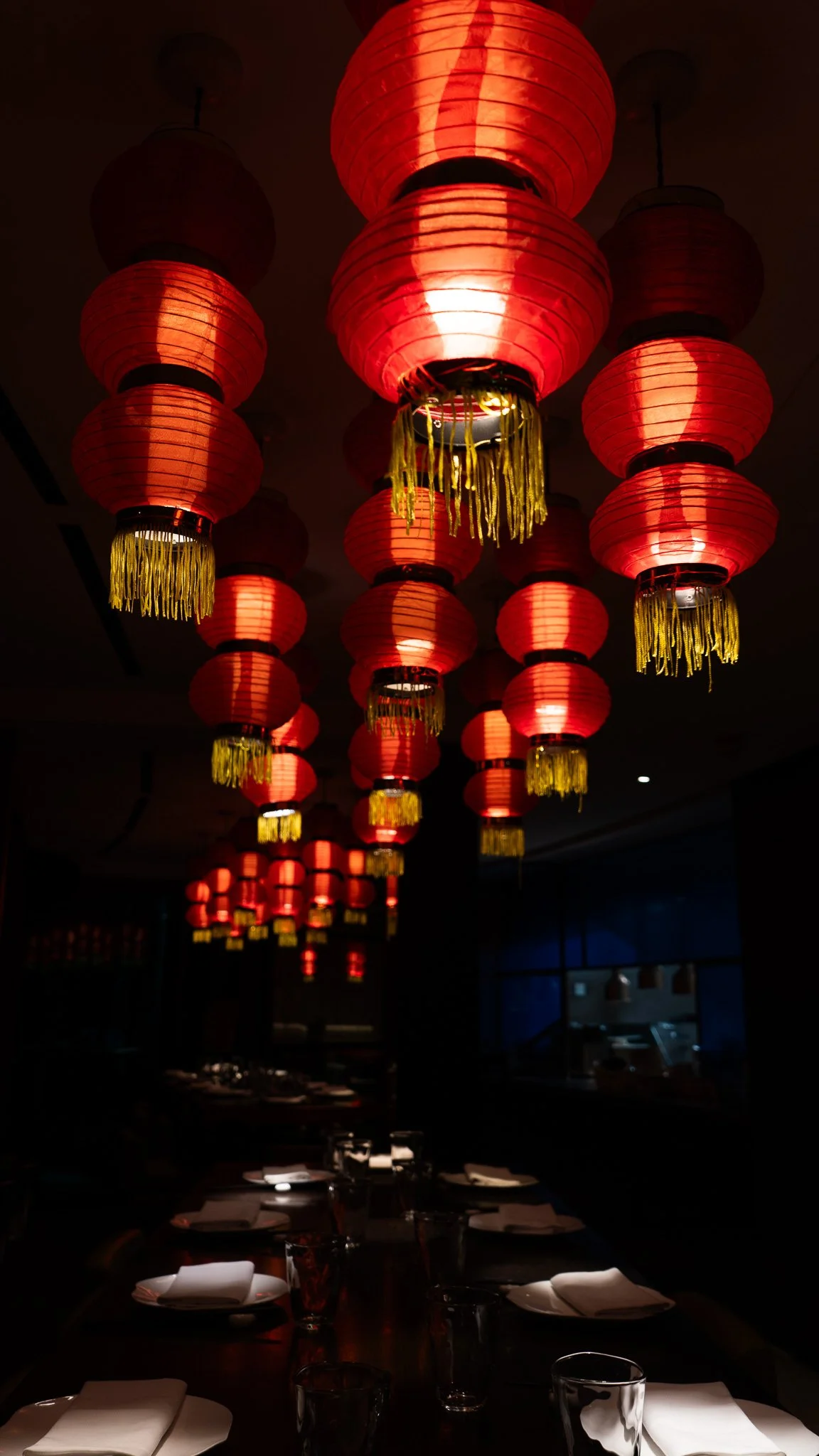 Red paper lanterns hanging from the ceiling in a dimly lit dining area, creating an illuminated pathway above a table set with white napkins and glasses.
