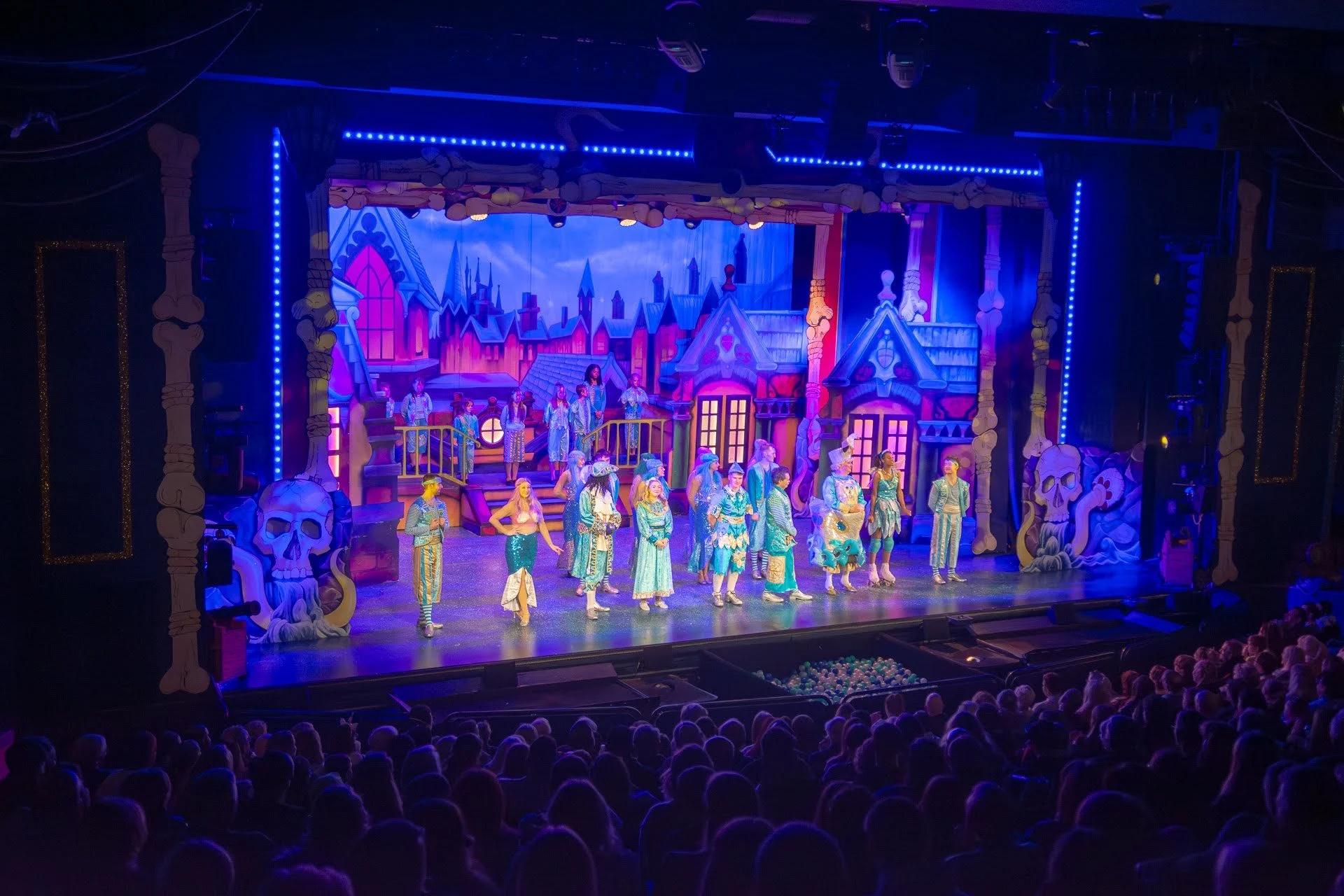 Theater stage with actors dressed in colorful costumes performing a musical, backdrop of a castle with tall spires, decorated with bones and skulls, audience watching.