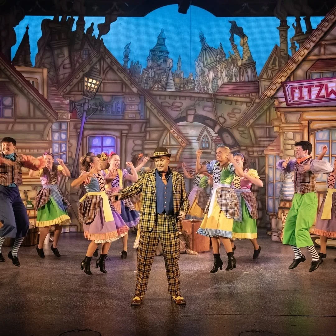 A musical theatre stage scene with a man in a plaid yellow and brown suit and hat, surrounded by male and female dancers in colorful 1950s-style costumes, performing in front of a backdrop of a cartoonish cityscape with old-fashioned buildings and si