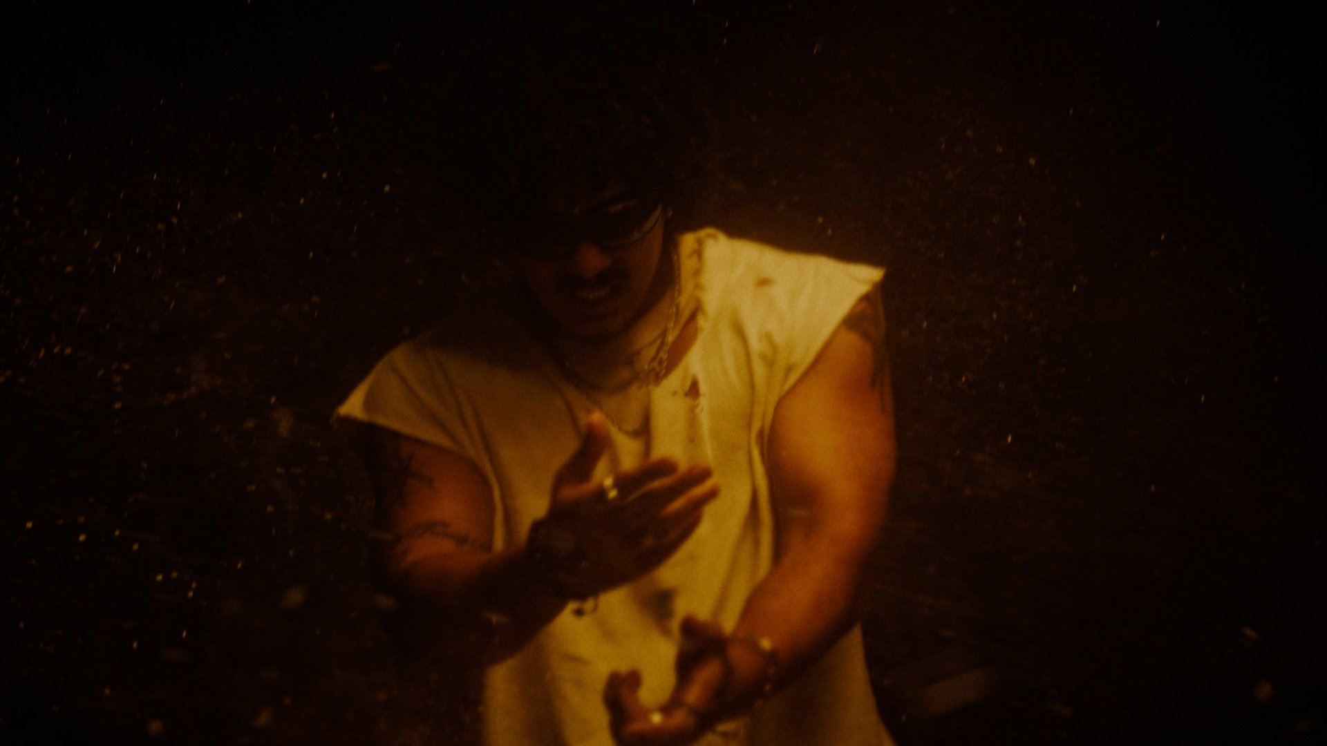 A person with glasses and long hair in a sleeveless shirt, standing in a dark environment with sparks or glowing particles around them.