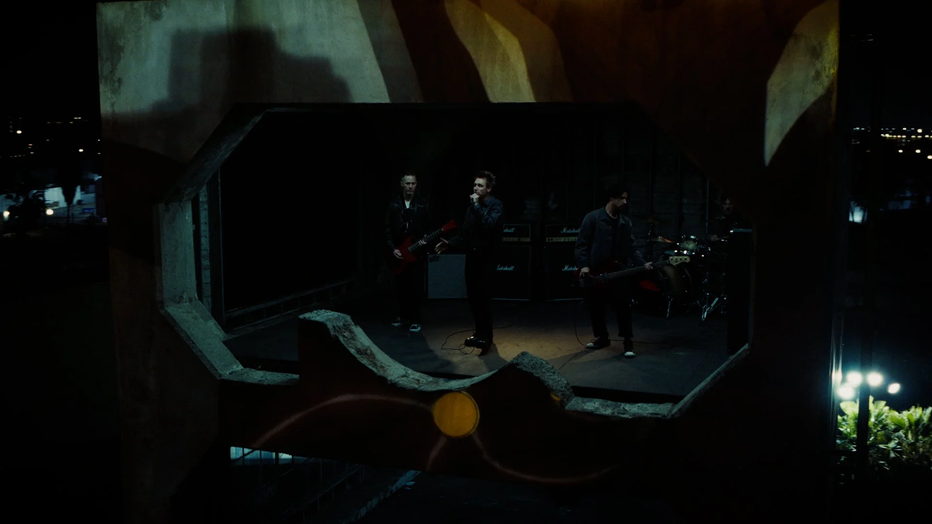 A band performing on an outdoor stage at night, viewed through a large concrete octagonal structure with a cityscape in the background. The band members are playing guitars and drums, with amplifiers behind them.