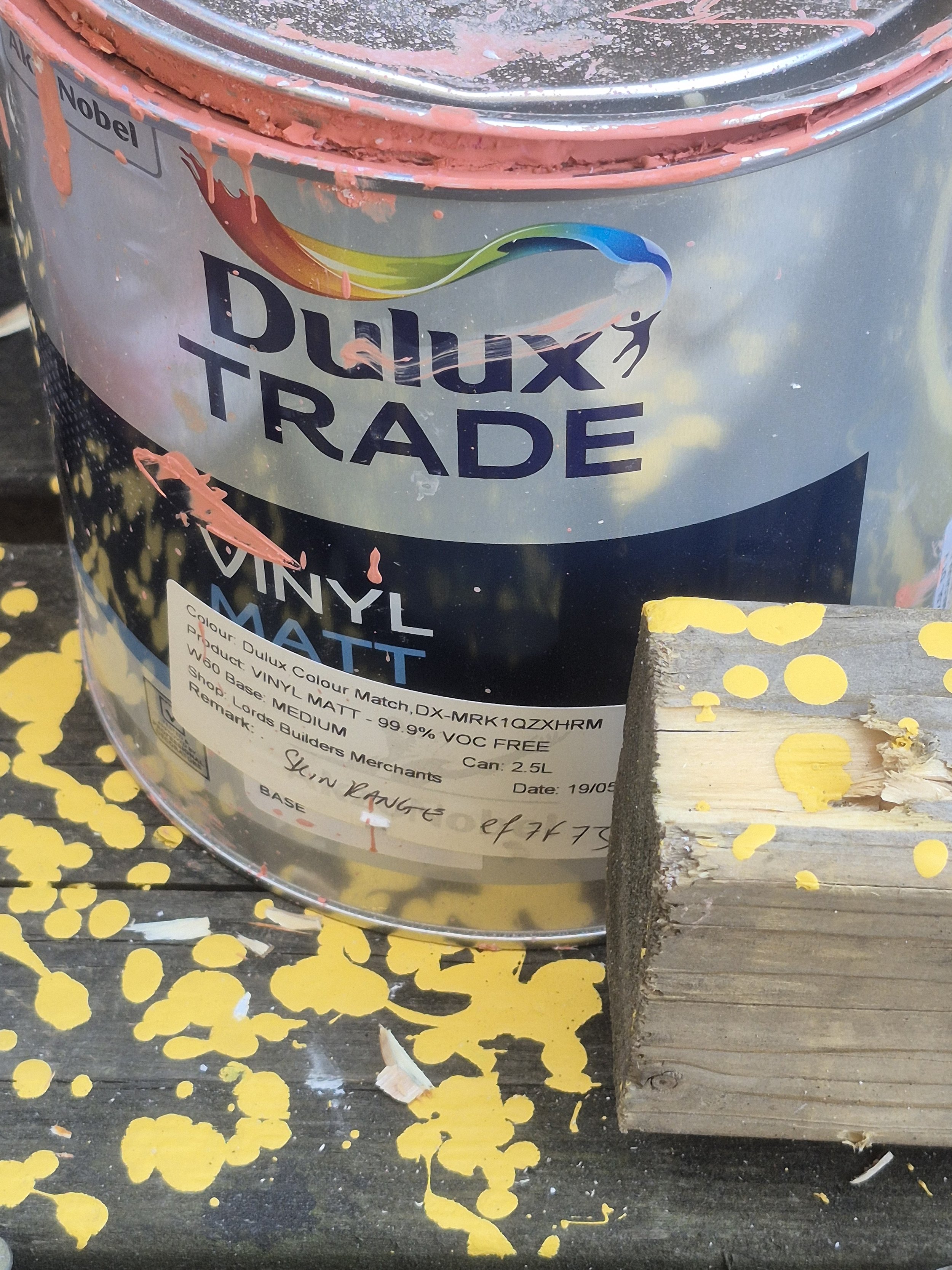 Close-up of a paint can labeled Dulux Trade, with pink paint smudged on the lid and sides, placed on a wooden surface with yellow painted spots.