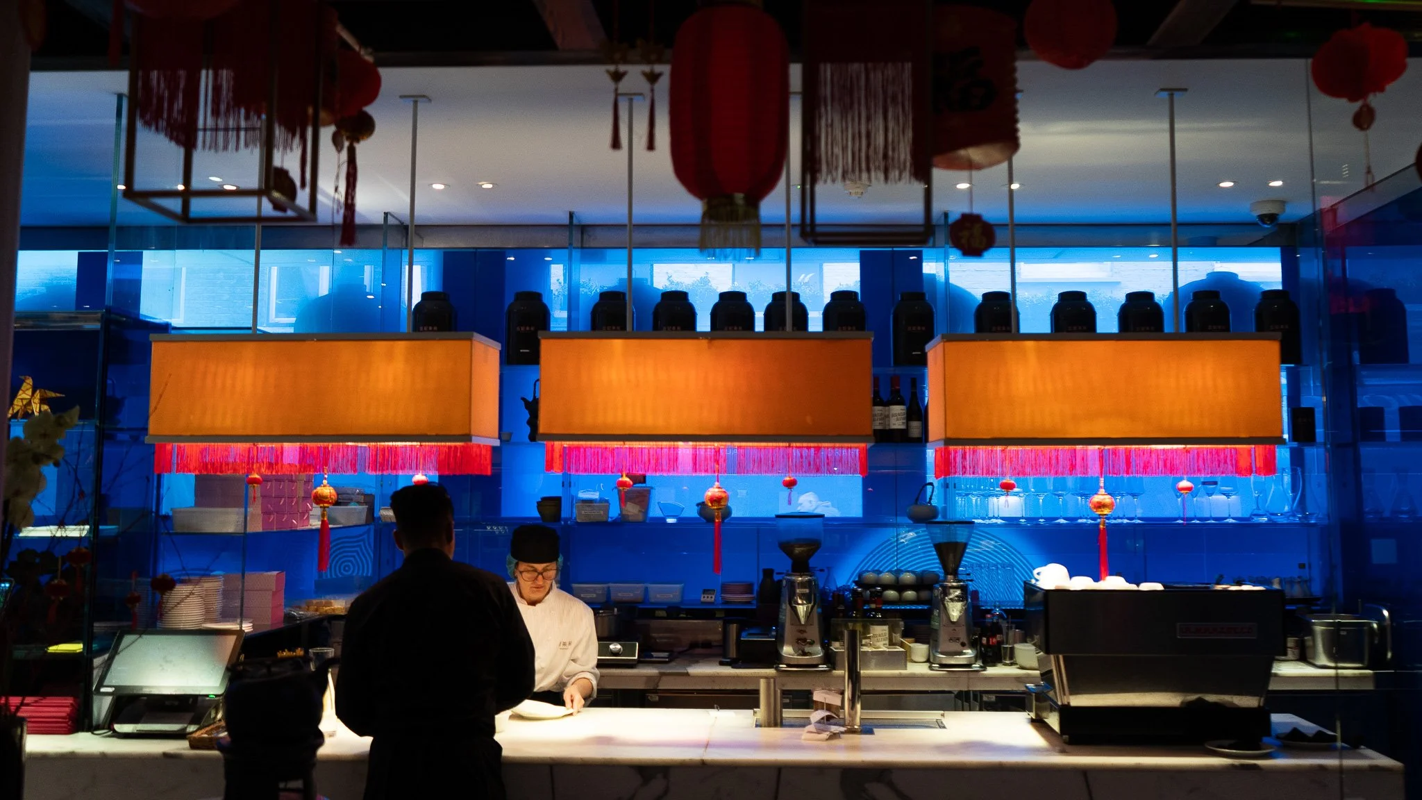 The image shows a restaurant or food service counter decorated with red lanterns and ornaments, featuring two chefs preparing food, with various containers and equipment behind them, and a blue-lit background with large windows.