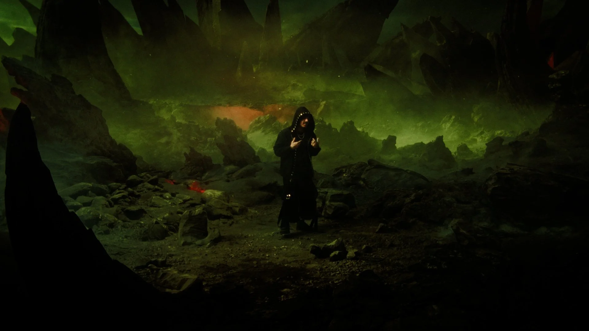 A person dressed in black with a hood and chains is standing on rocky terrain inside a cave illuminated by greenish light, with a dark figure visible on the left side.
