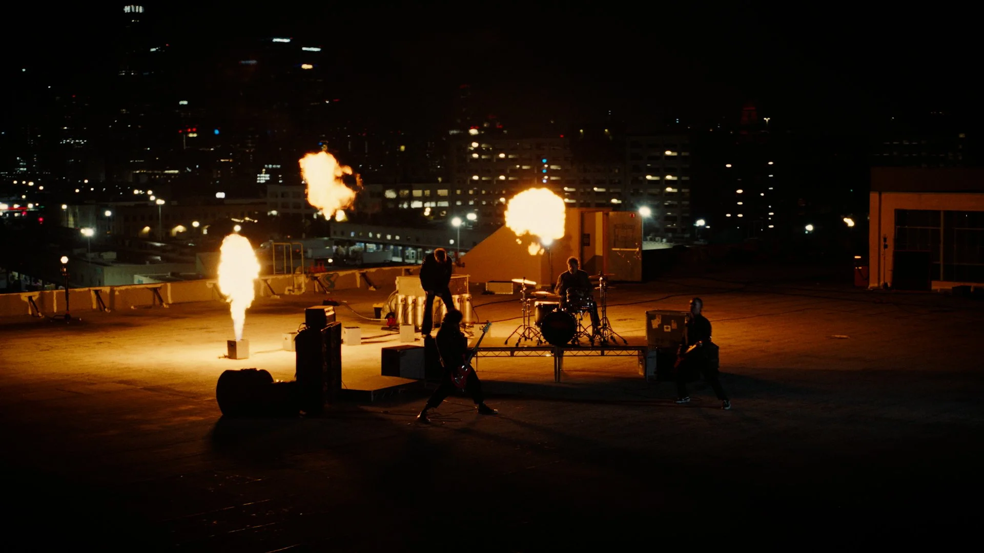 A band performing on a rooftop at night with flames and fire effects, city buildings in the background.
