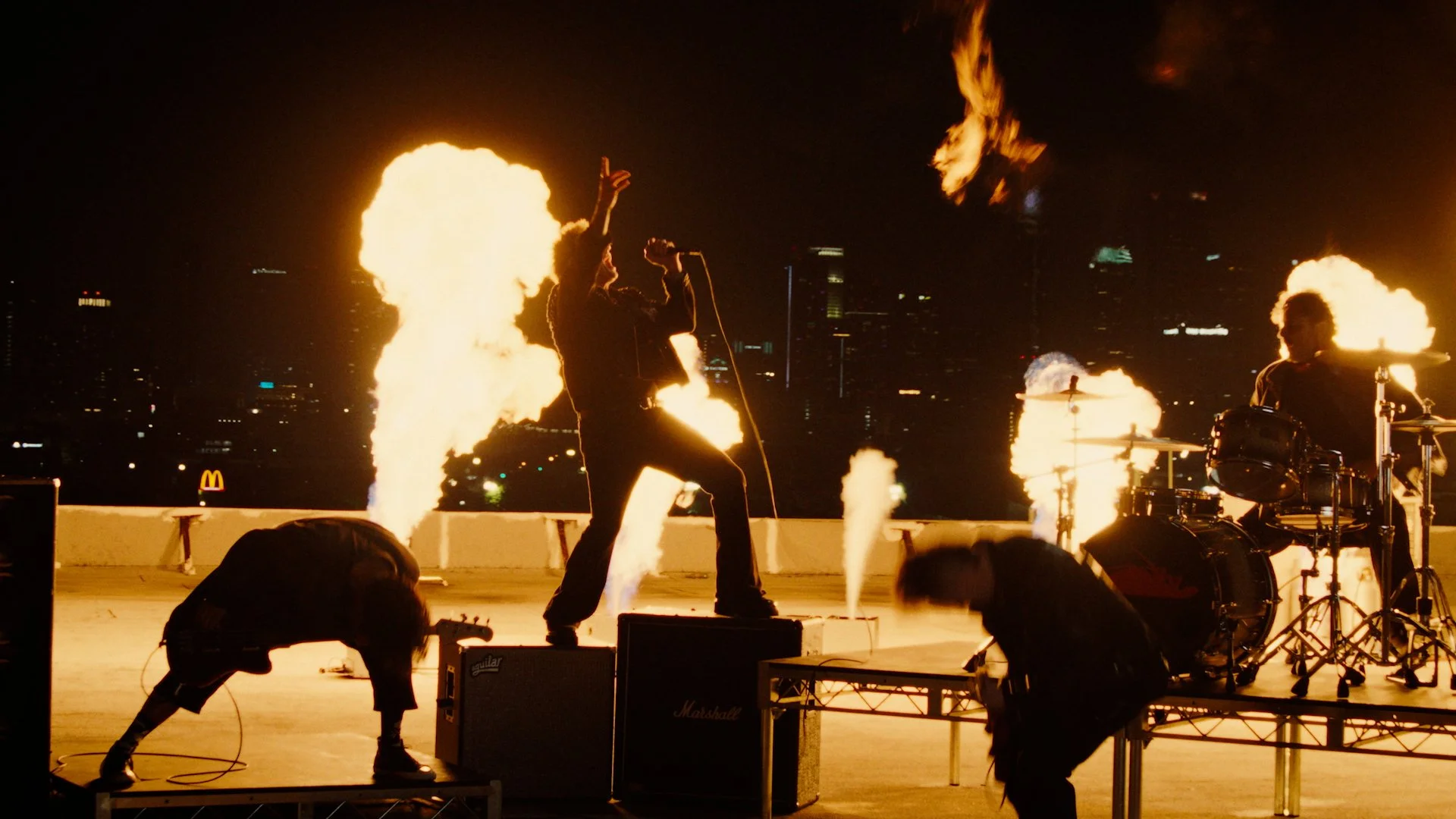 Band performing on a rooftop at night with flames and fire effects, city skyline in background.