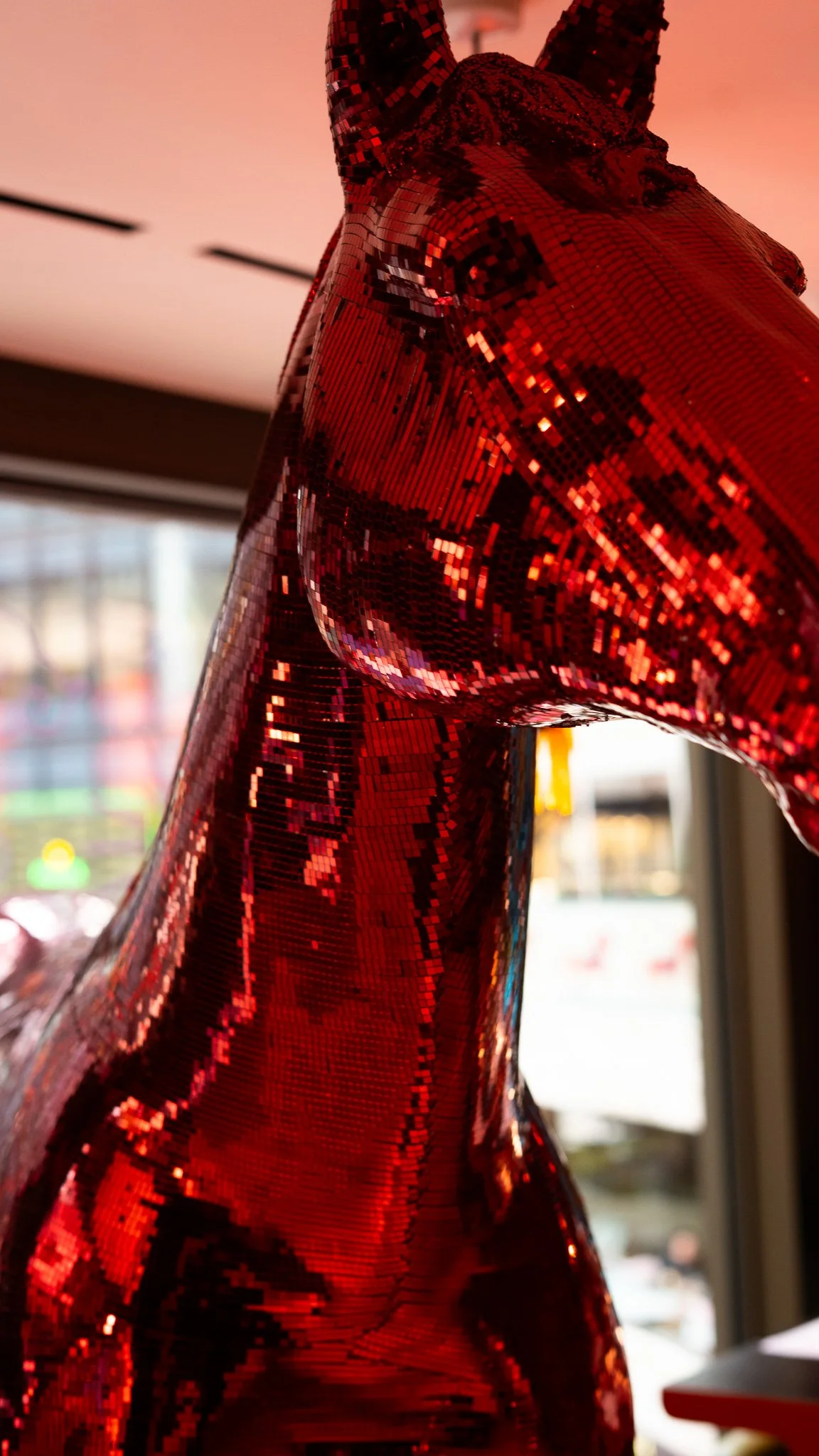 Close-up of a shiny red, pixelated, devil-like sculpture with horns, set indoors with blurred background.